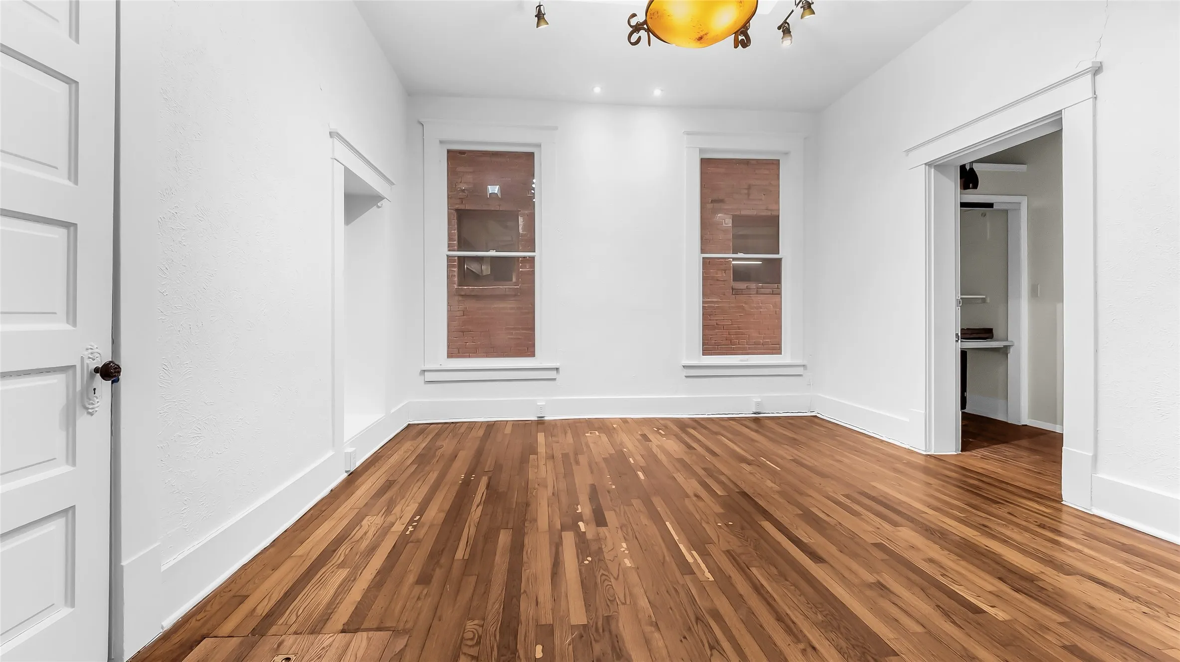 Hardwood flooring, white painted walls, two large windows, overhead light fixture, and panel door