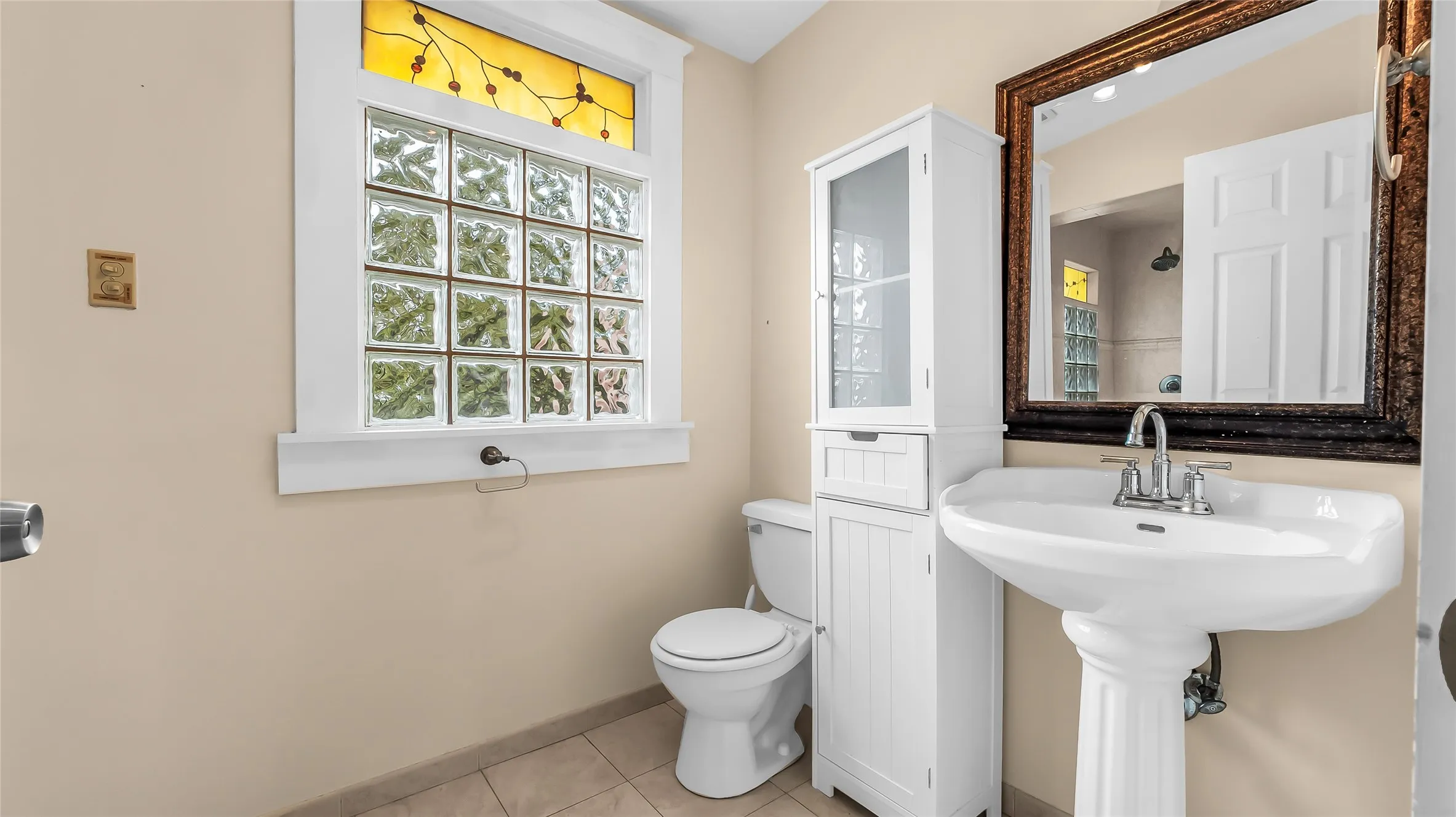 Pedestal sink with chrome fixtures, tiled flooring, glass block window with stained glass transom, and a tall storage cabinet