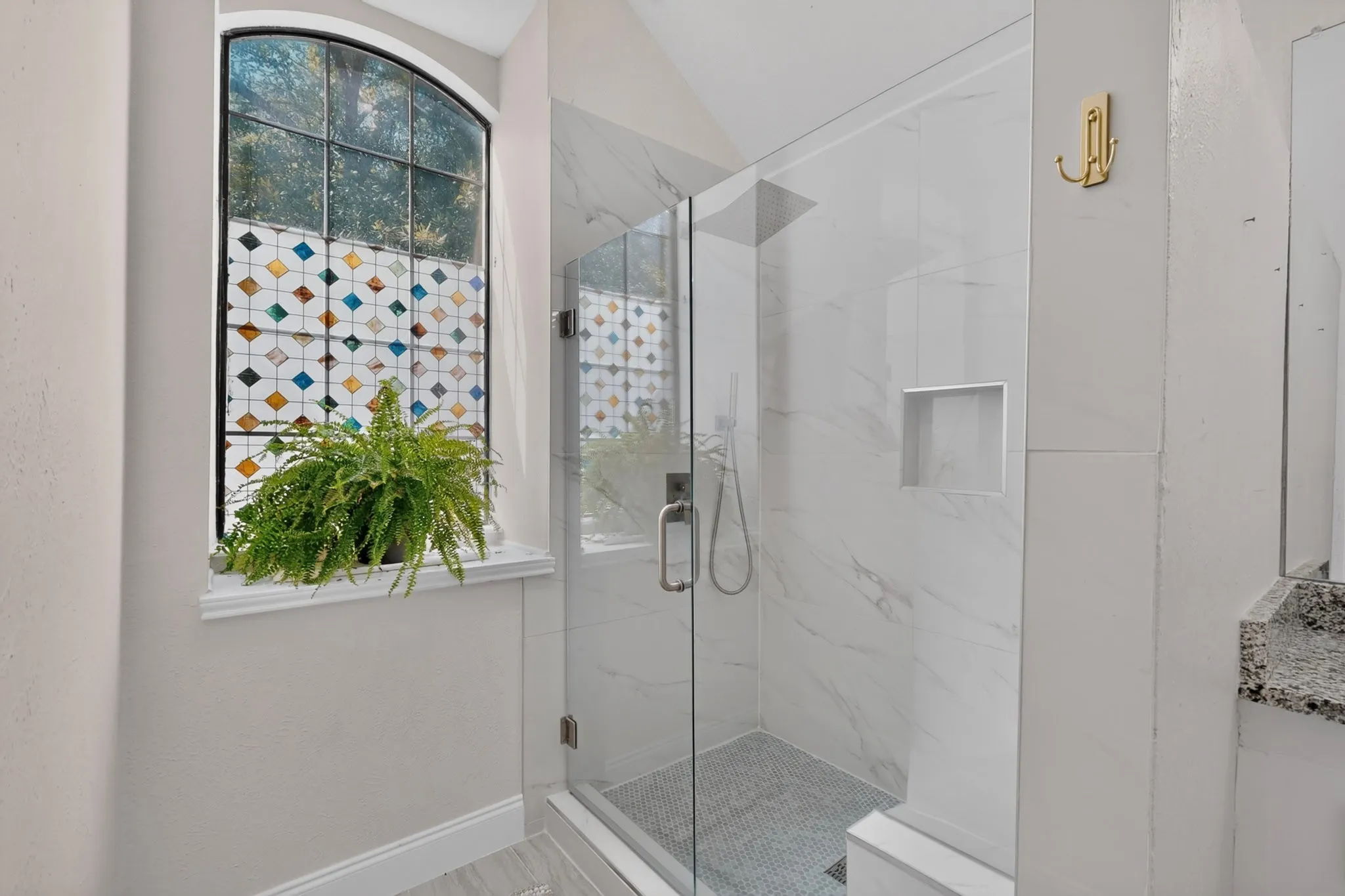 Arched window with decorative film, glass-enclosed shower, white marble-look shower surround, mosaic tile shower floor, and built-in shower niche