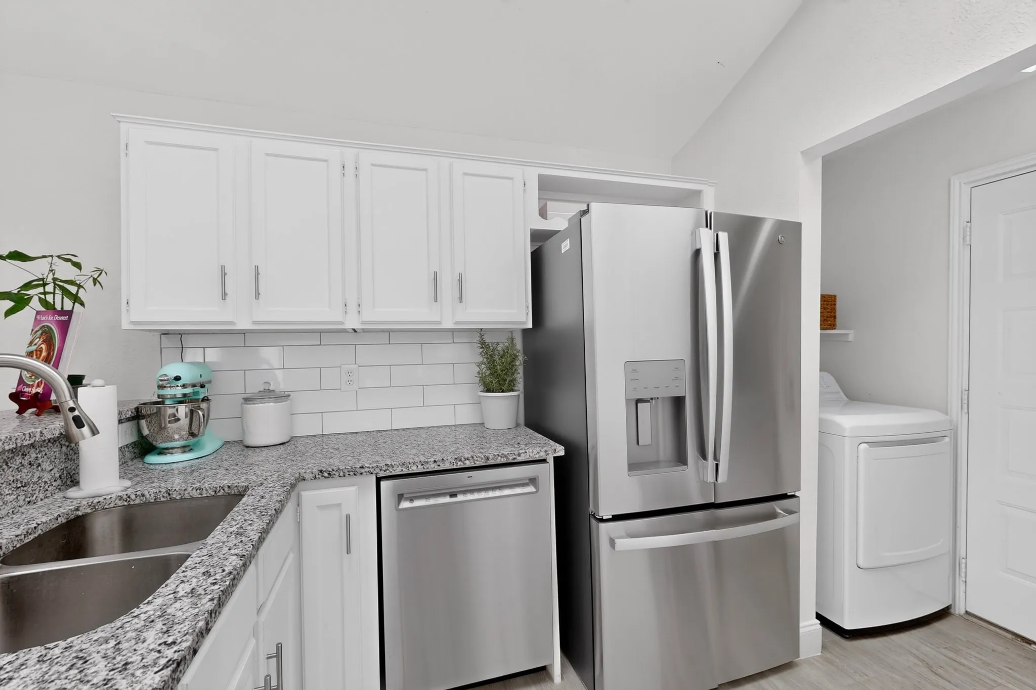 White cabinetry with stainless steel hardware, L-shaped speckled countertop, dual basin stainless steel sink, subway tile backsplash, and stainless steel appliances