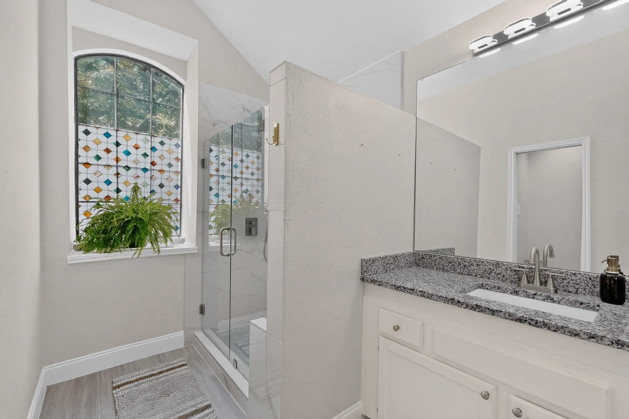 Arched window with decorative film, glass-enclosed shower, white vanity with granite-style countertop, and wood-finish flooring