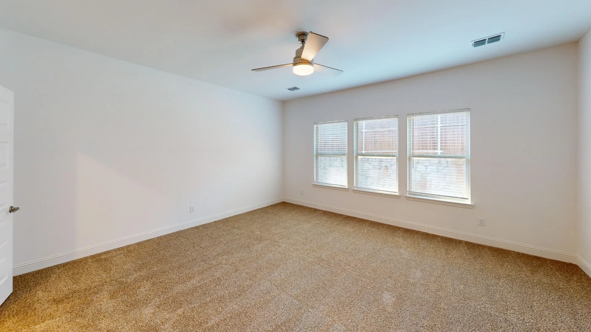 Neutral wall color, light brown carpet, three windows with blinds, ceiling fan with integrated lighting, and white door with silver hardware