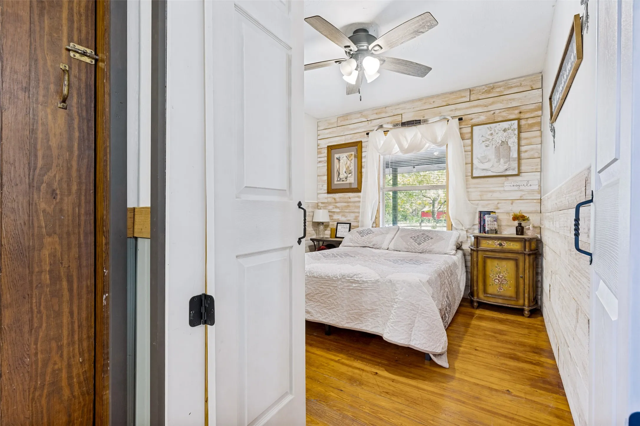 Wood-finish flooring, white shiplap accent wall, ceiling fan with integrated lighting, white paneled interior doors, and a window with white trim