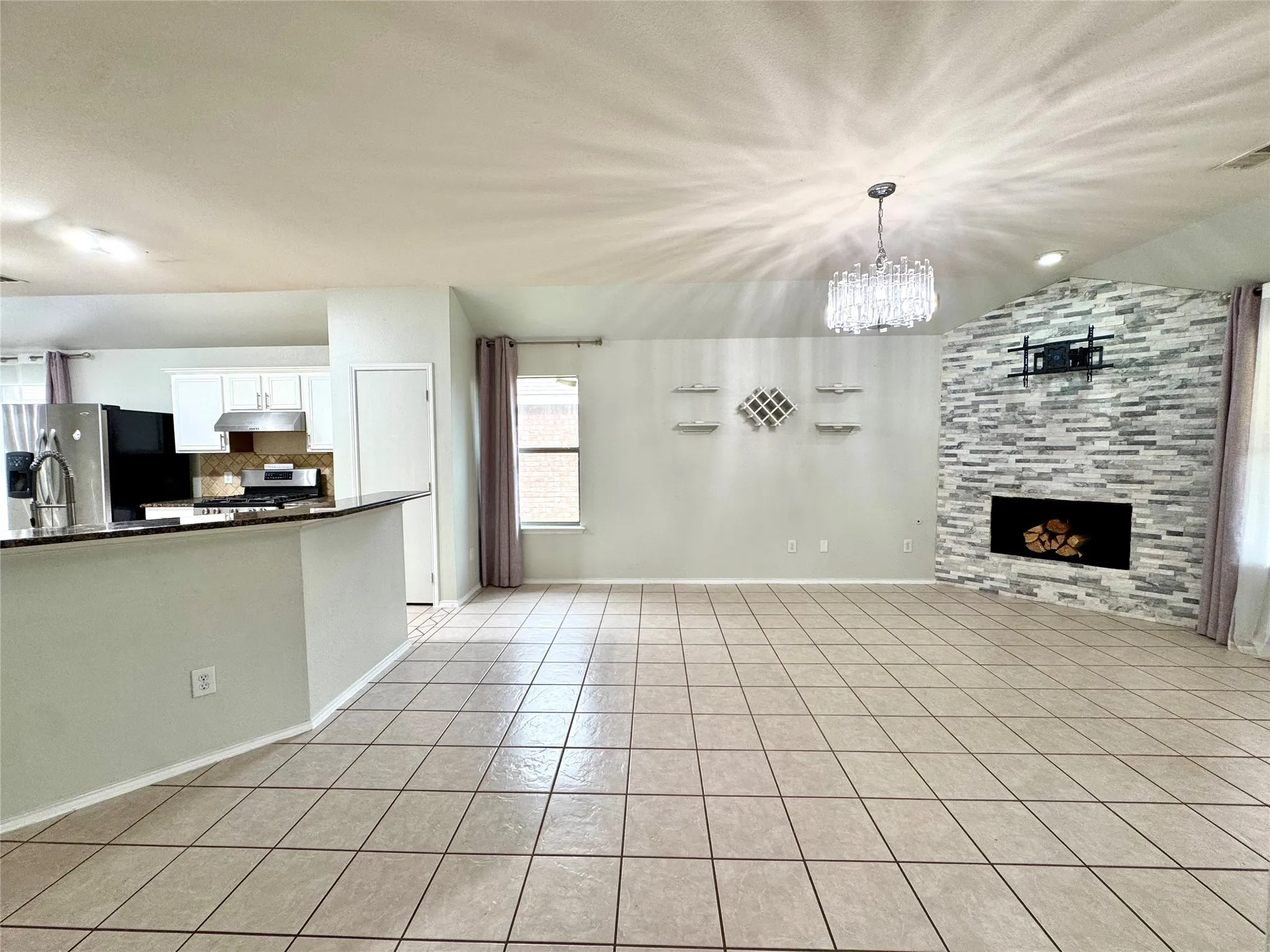 Tiled flooring throughout, vaulted ceiling with a crystal chandelier, stone-clad fireplace, built-in shelving, and a kitchen pass-through with a breakfast bar