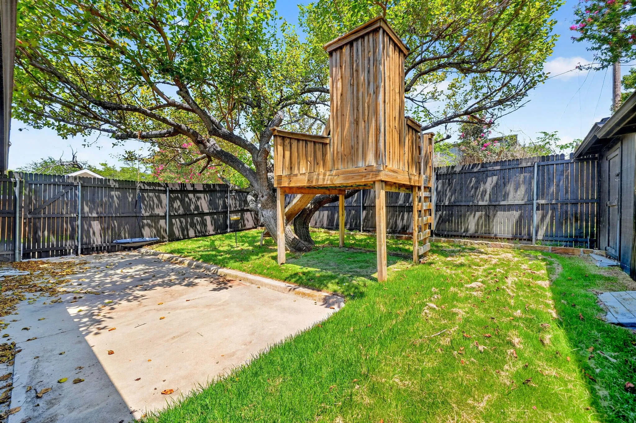 Grassed yard with a large shade tree, a wood-built playhouse, and a concrete pad