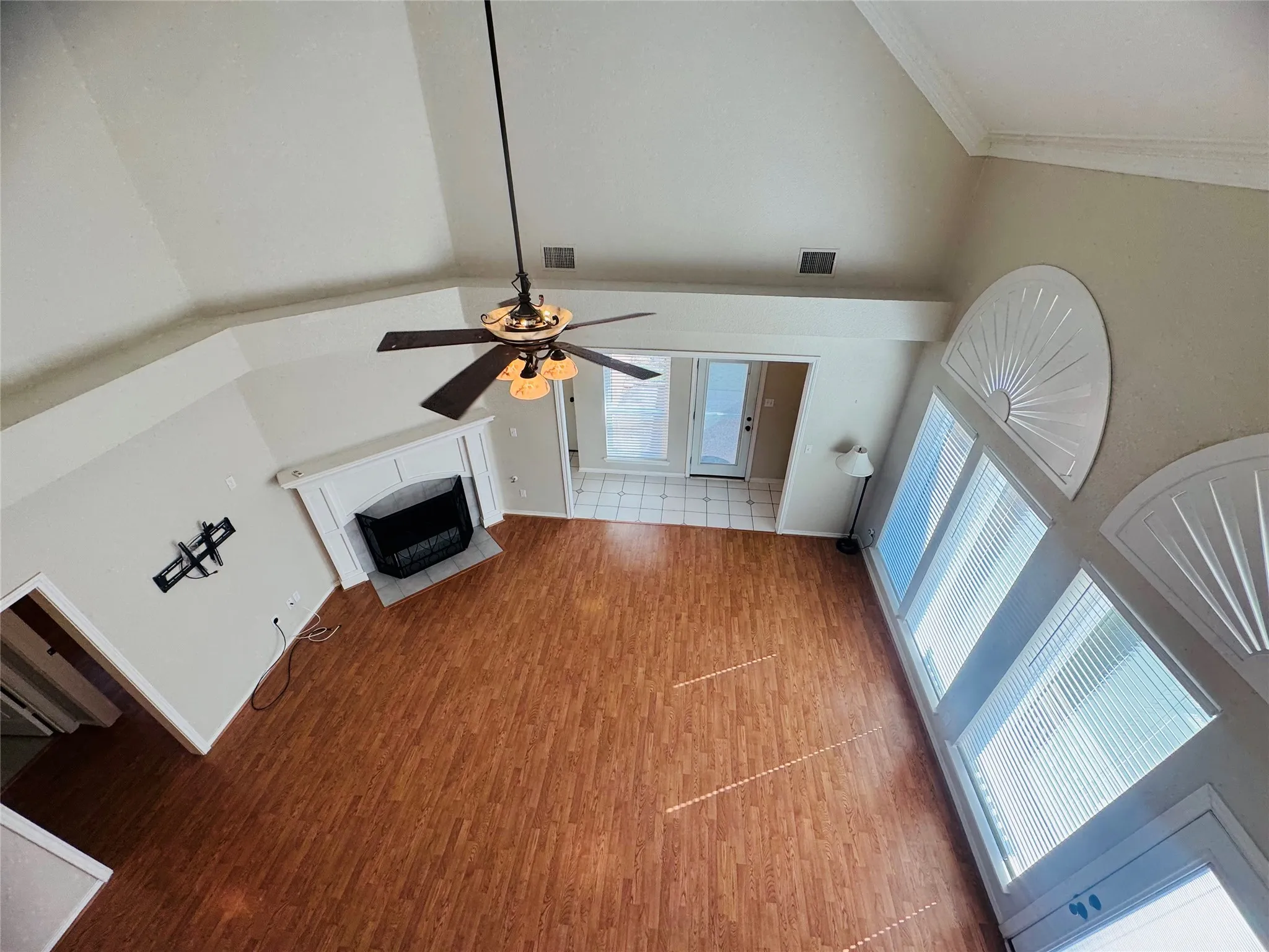 Vaulted ceiling, wood-finish flooring, white mantel fireplace, ceiling fan with integrated lighting, and multiple windows with blinds