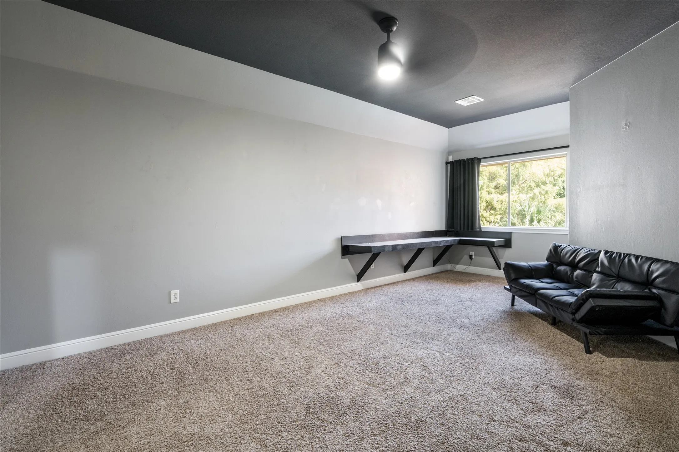 Recessed ceiling with integrated lighting, built-in corner desk, window with dark curtains, and neutral carpet flooring