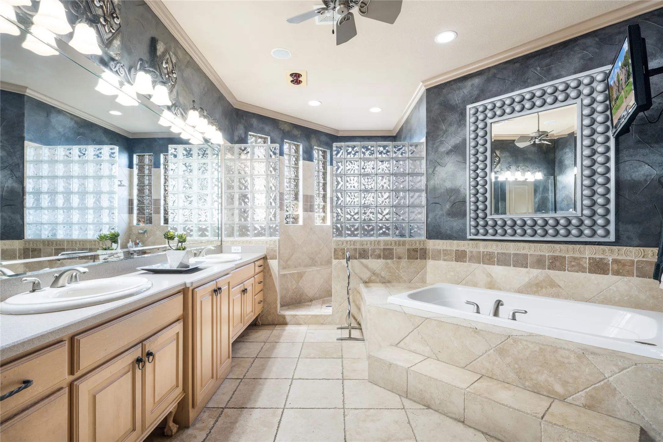 Dual sink vanity with light wood-finish cabinetry, built-in bathtub with tiled surround, and glass block shower enclosure