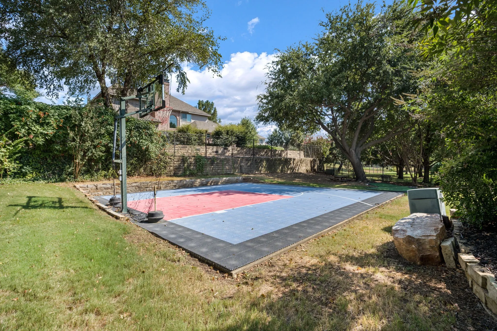 Hardscaped sport court with basketball hoop, surrounded by established greenery and a stone retaining wall