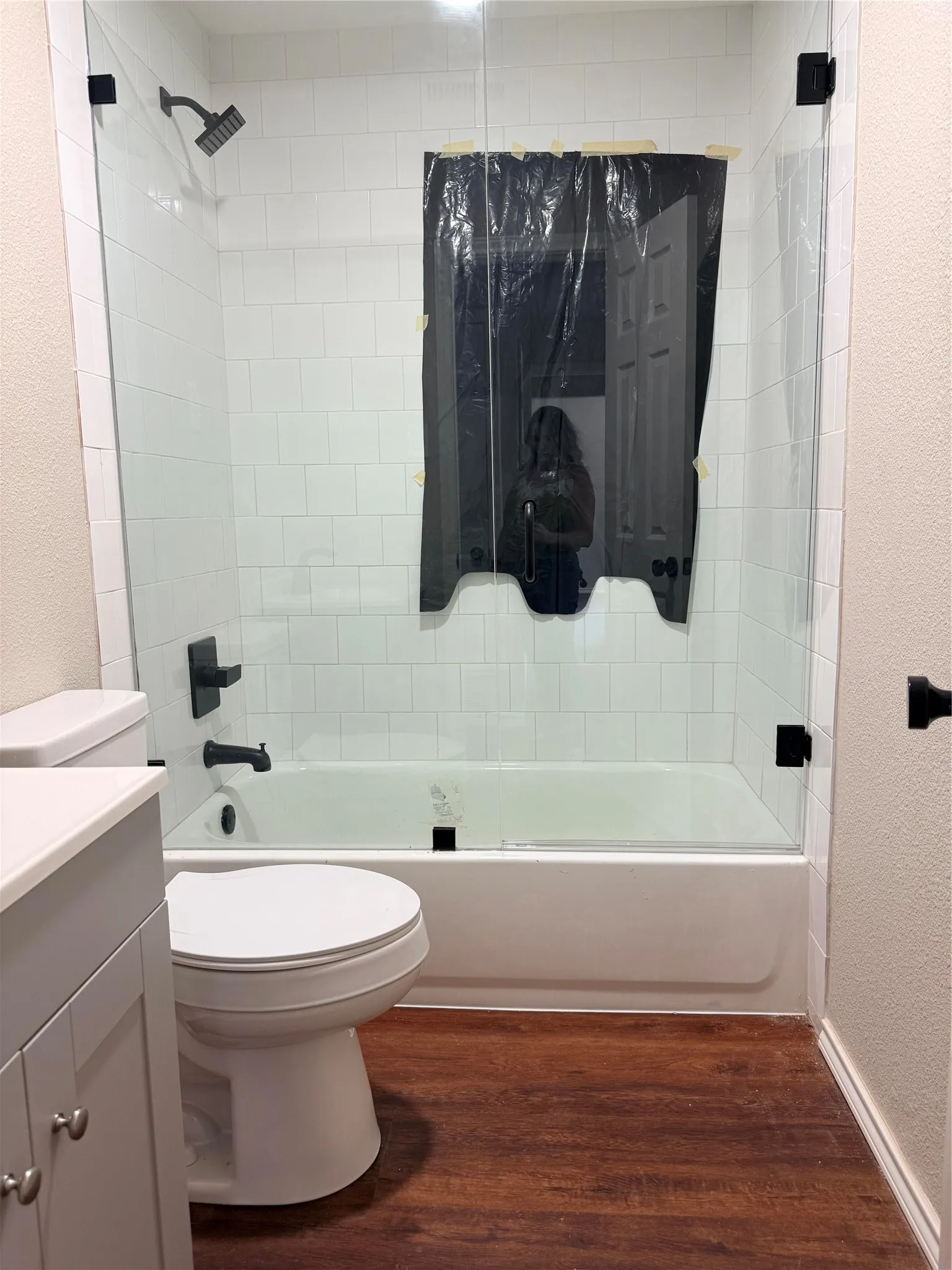 Bathtub and shower combination with clear glass enclosure and matte black fixtures, white subway tile surround, wood-finish flooring, and light-colored vanity with white countertop