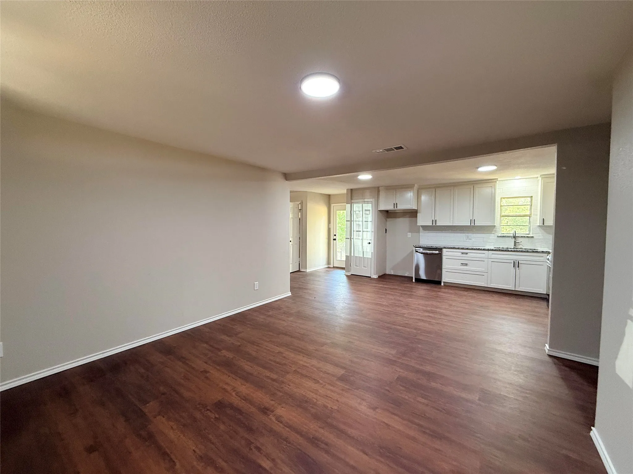 Open-concept living area with wood-finish flooring, recessed lighting, and a kitchen featuring white cabinetry, a stainless steel dishwasher, and a window above the sink