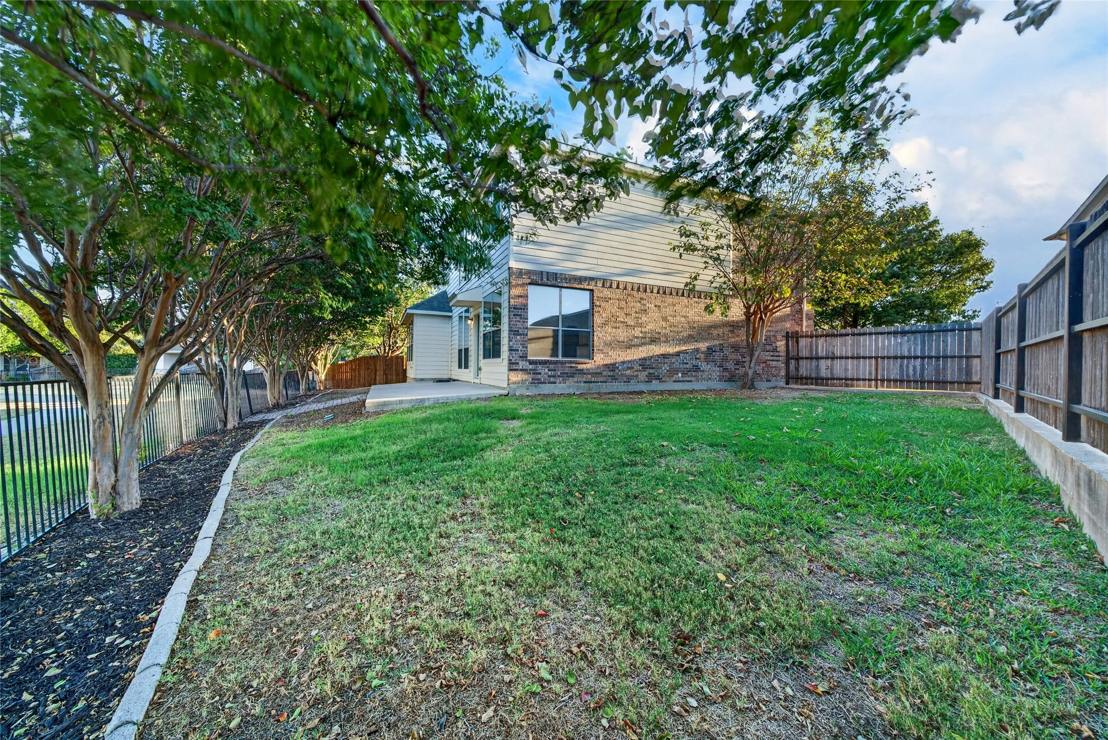Expansive backyard featuring a concrete patio, brick and siding exterior, established trees, and perimeter fencing