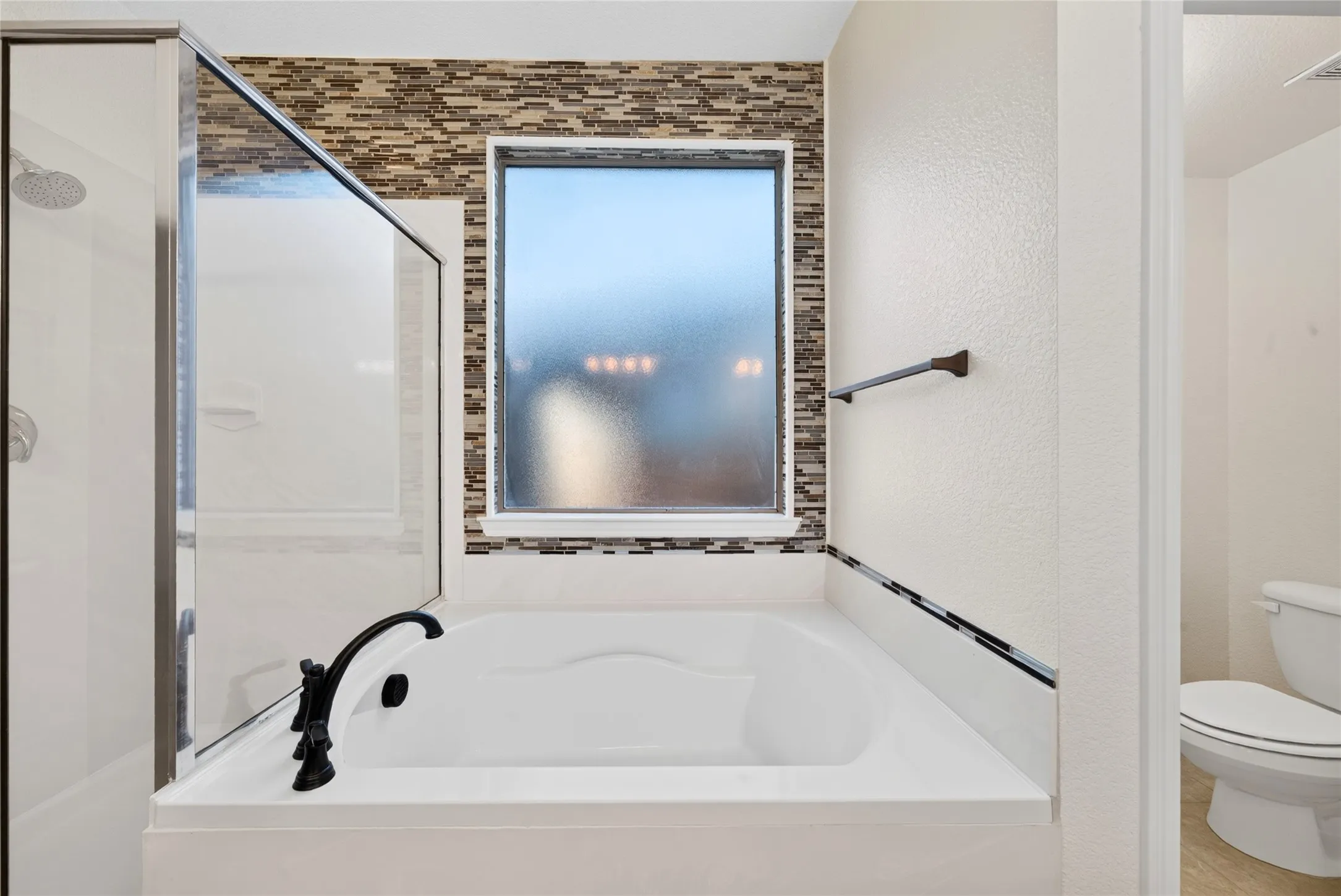 Soaking tub with black fixtures, framed window with privacy glass, tiled accent wall, and glass-enclosed shower