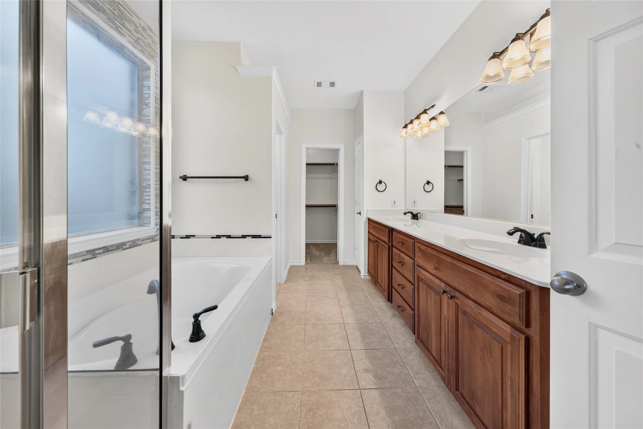 Dual vanity with wood-finish cabinetry, ceramic tile flooring, built-in bathtub, and an enclosed shower with a mosaic tile accent