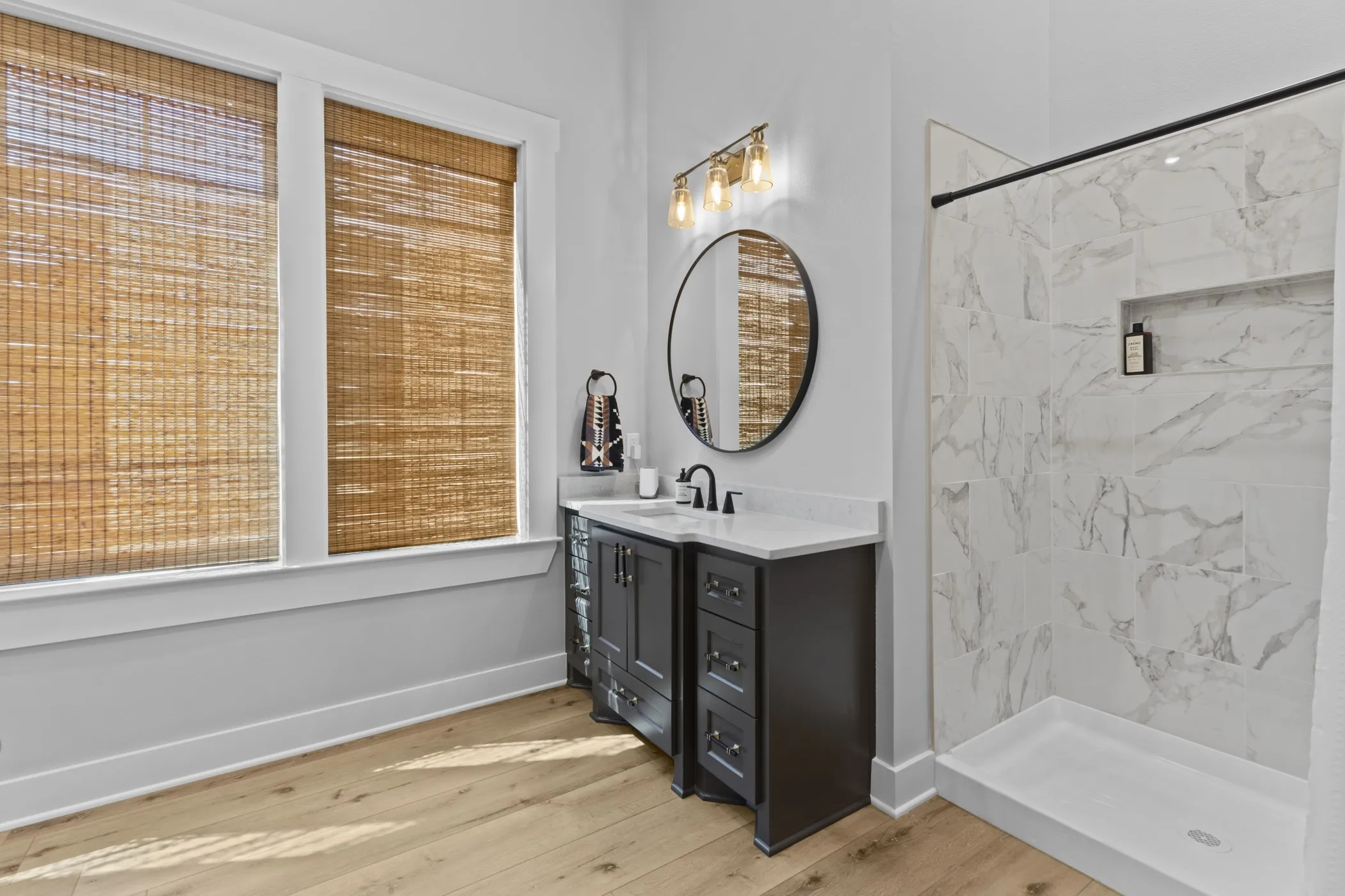 Wood-finish flooring, dark wood vanity with white countertop, round wall-mounted mirror, walk-in shower with tile surround, and dual windows with natural woven shades