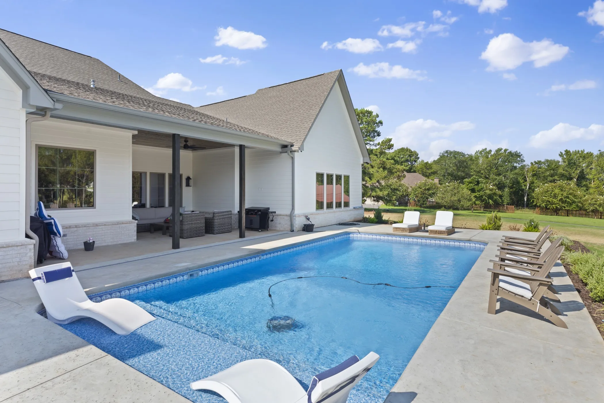 Outdoor pool with an integrated sun shelf, concrete decking, and a covered patio featuring dark columns and a shingle roof