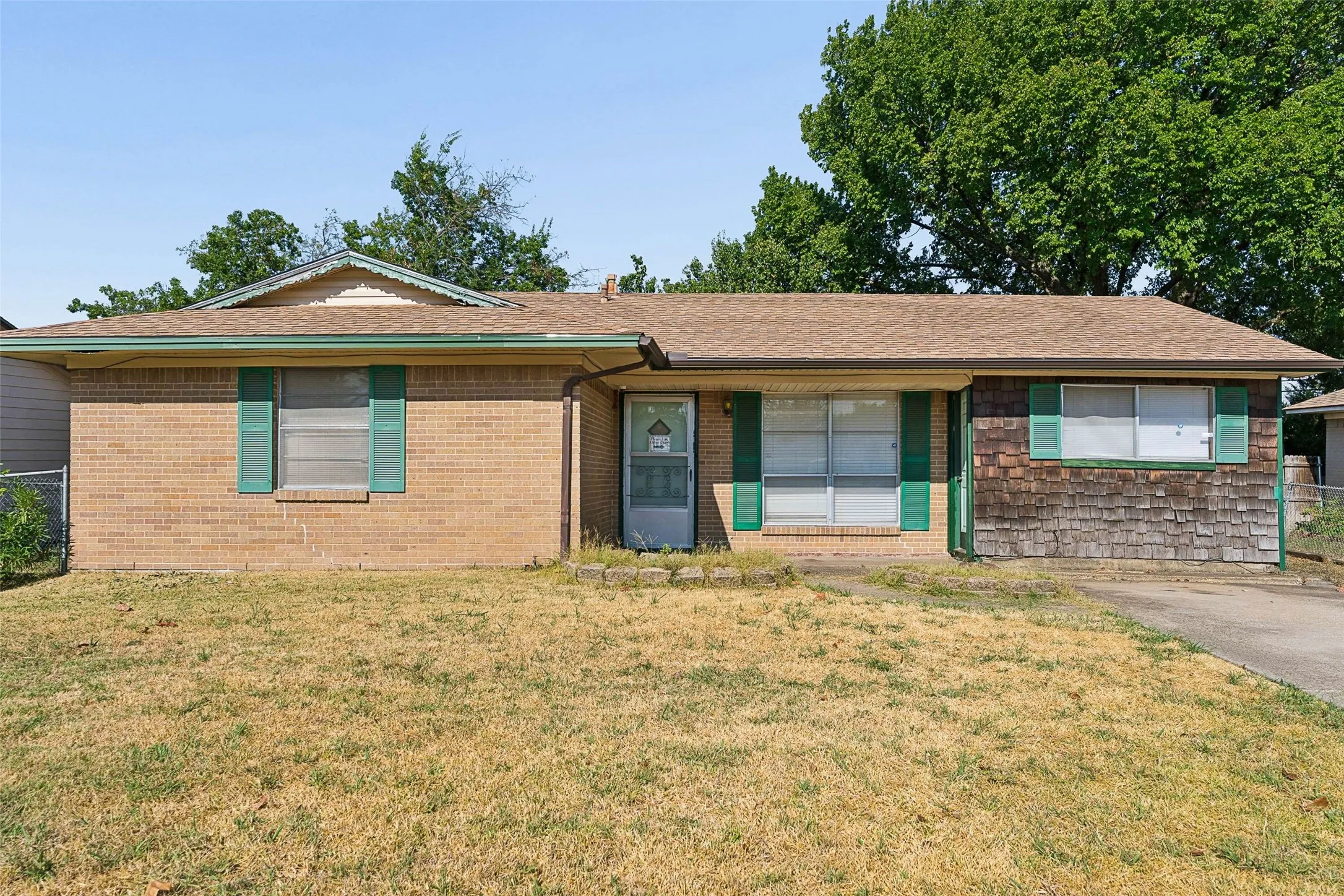 Brick and shingle exterior with green window shutters, a brown shingle roof, and a storm door entry