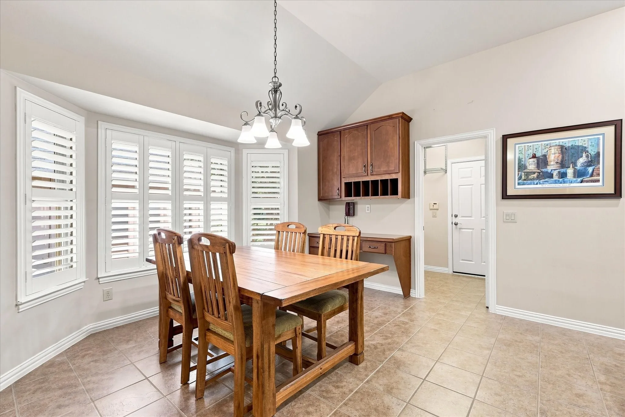 Vaulted ceiling with an chandelier, bay window with plantation shutters, built-in desk with cabinetry, and tile flooring