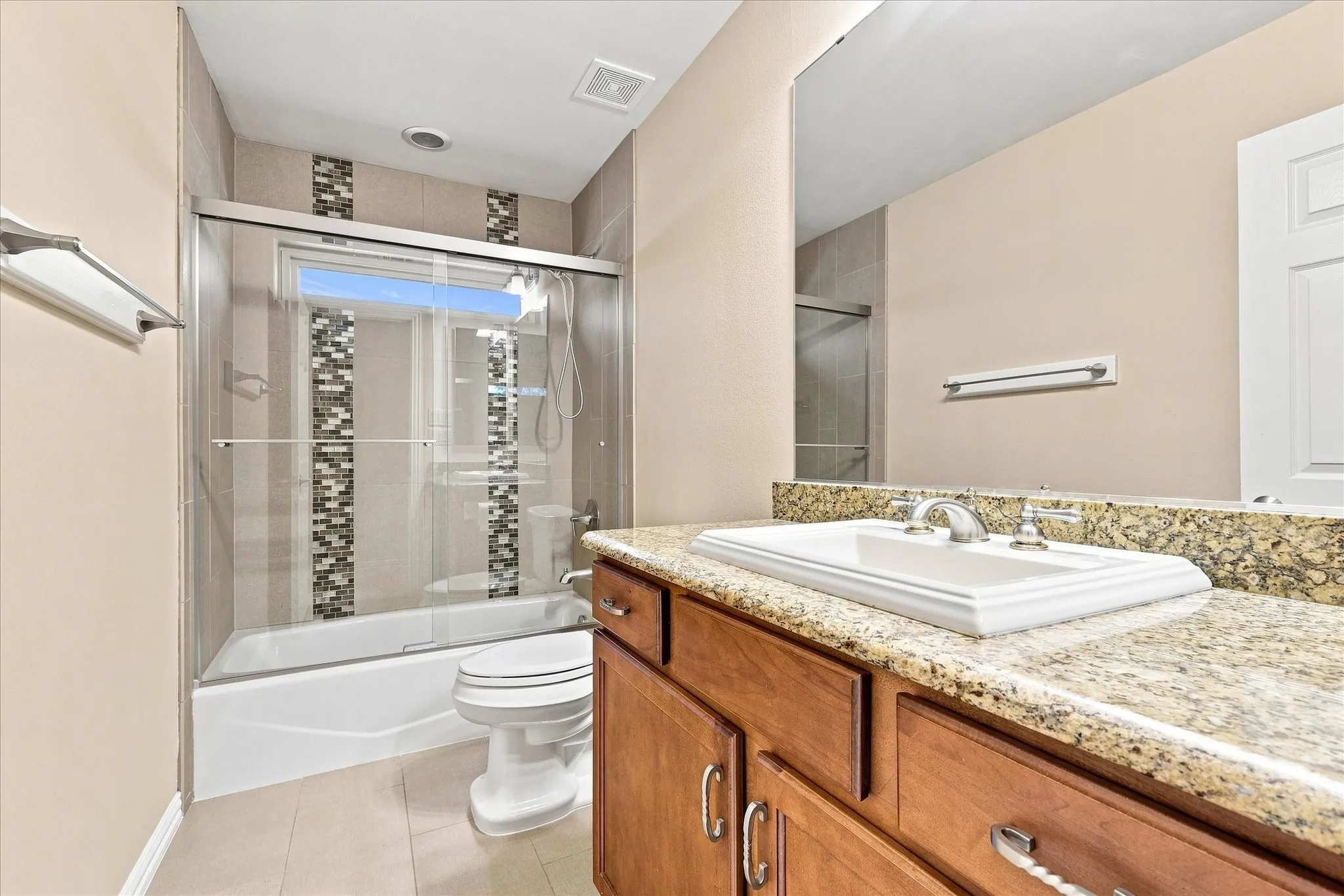 Bathroom featuring a wood-finish vanity with a granite countertop, tub with sliding glass enclosure, and tiled shower surround with accent mosaic