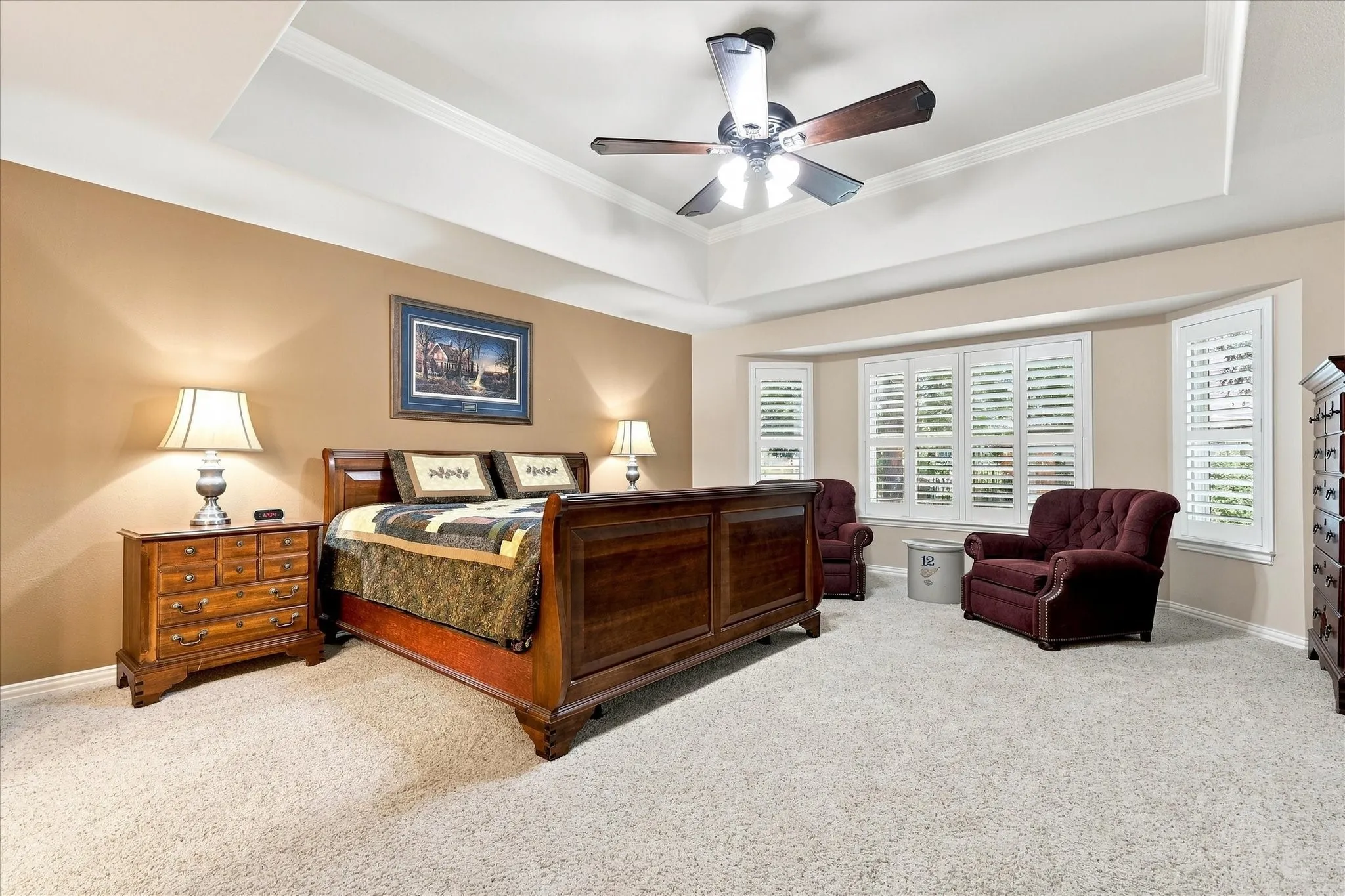 Tray ceiling with crown molding, ceiling fan with integrated lighting, bay window with plantation shutters, and light-toned carpeting