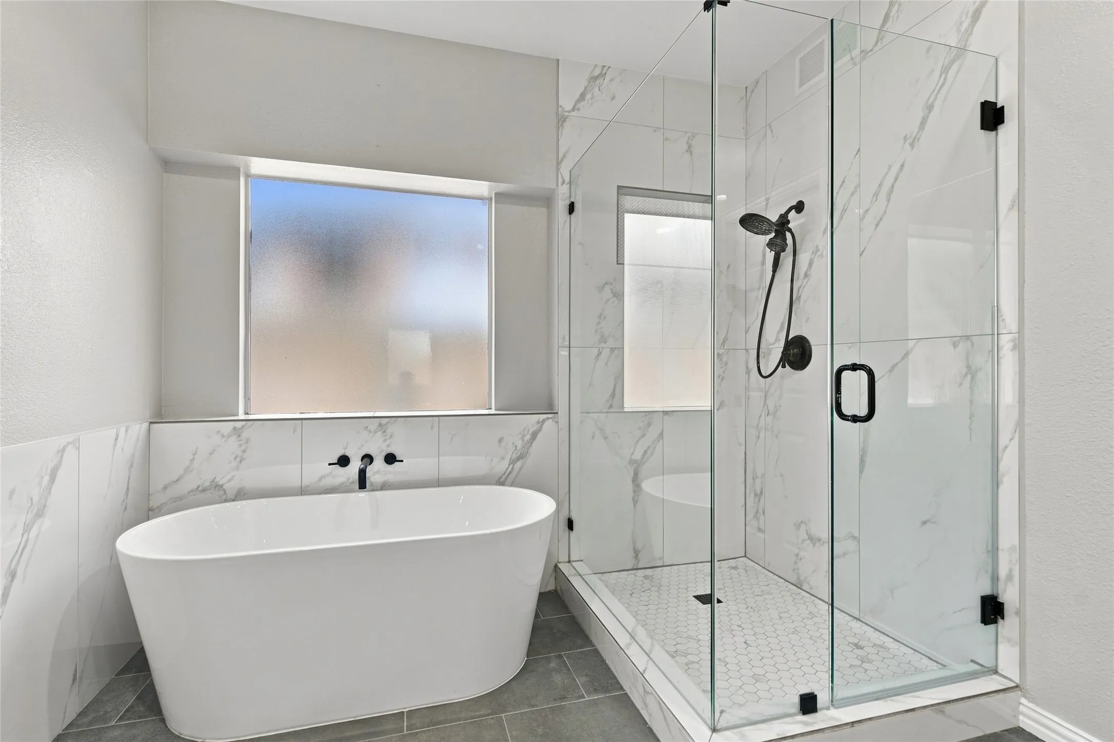 Freestanding soaking tub with wall-mounted matte black fixtures, marble-look tile surround, and a glass-enclosed shower with hexagon tile floor.