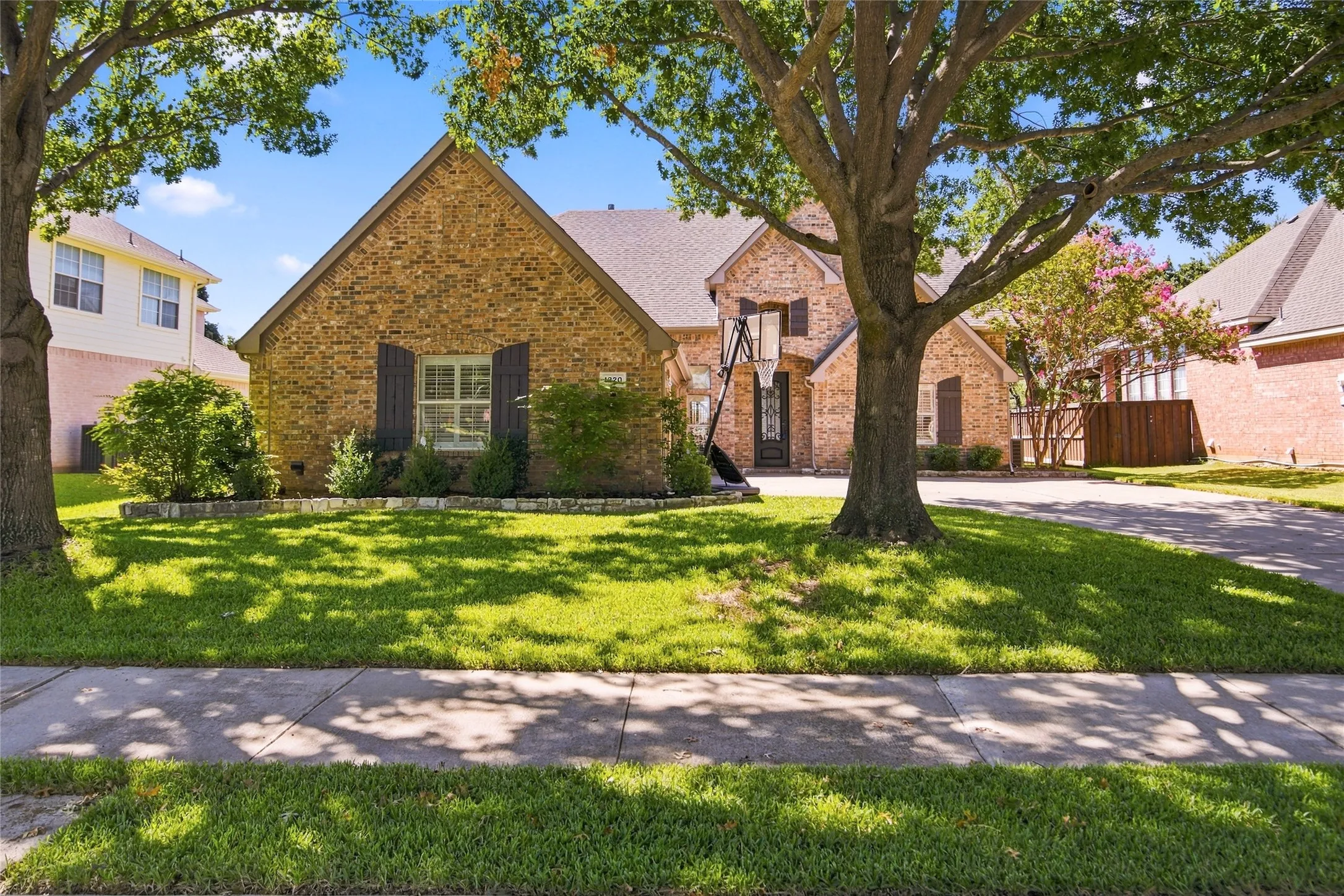 Brick exterior with contrasting dark window shutters, gabled roofline, mature trees, and a lush lawn