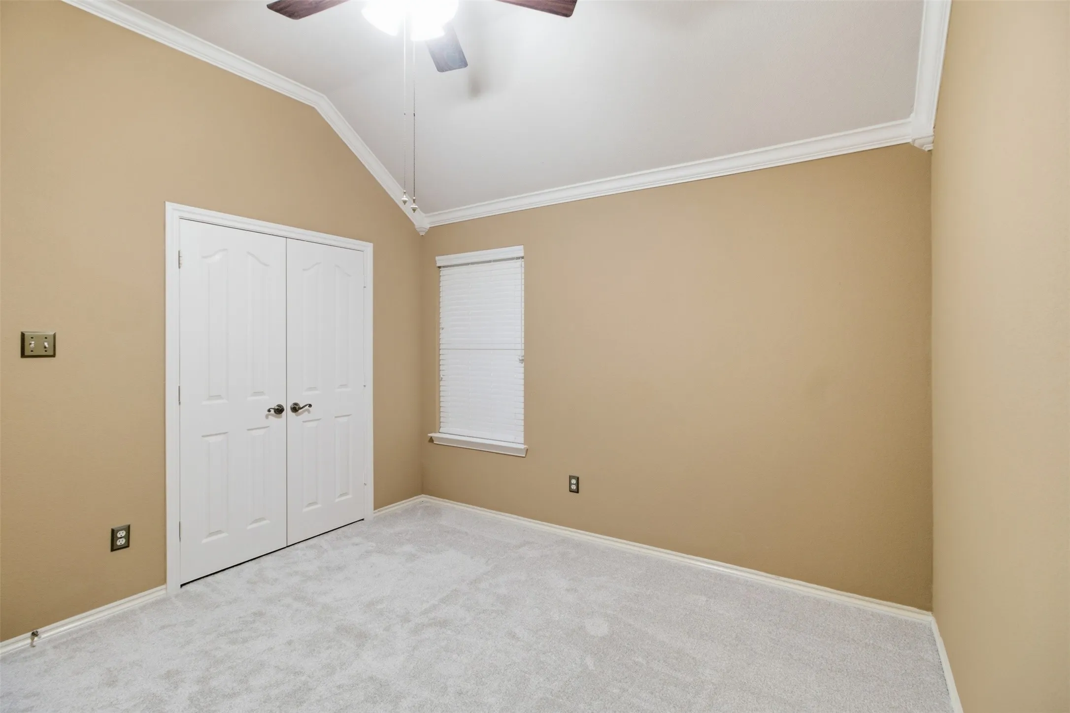 Vaulted ceiling with crown molding, double bifold doors, window with horizontal blinds, ceiling fan with light, and light-colored carpeting