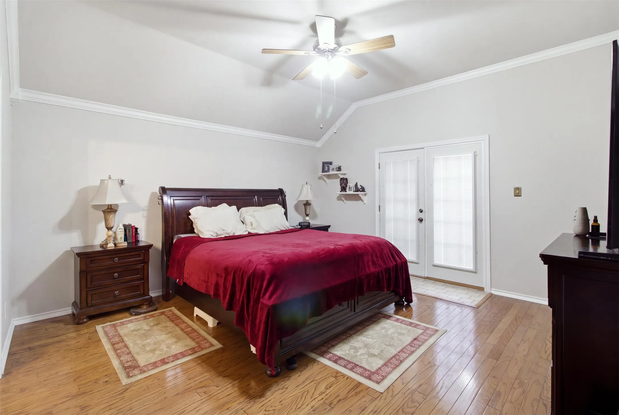 Vaulted ceiling with crown molding, wood-finish flooring, ceiling fan with light fixture, and double glass-paneled doors with blinds