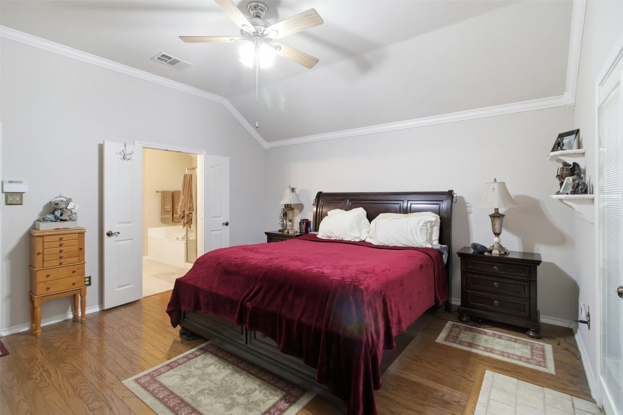 Vaulted ceiling with crown molding, wood-finish flooring, ceiling fan with integrated lighting, and double doors to a separate bathroom featuring a built-in tub