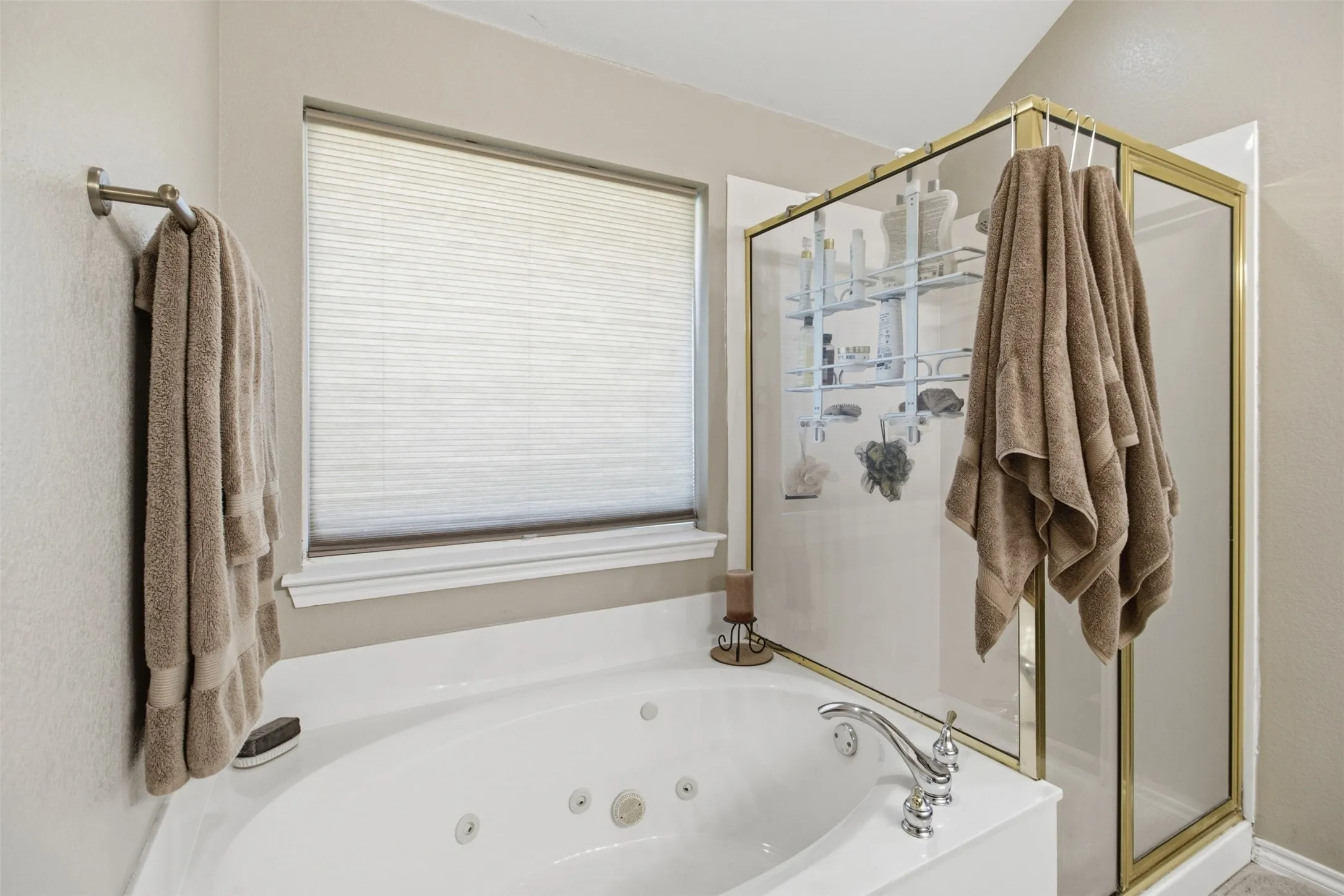 Jetted tub with chrome fixtures, an adjacent glass-enclosed shower with a gold-tone frame, and a window with blinds