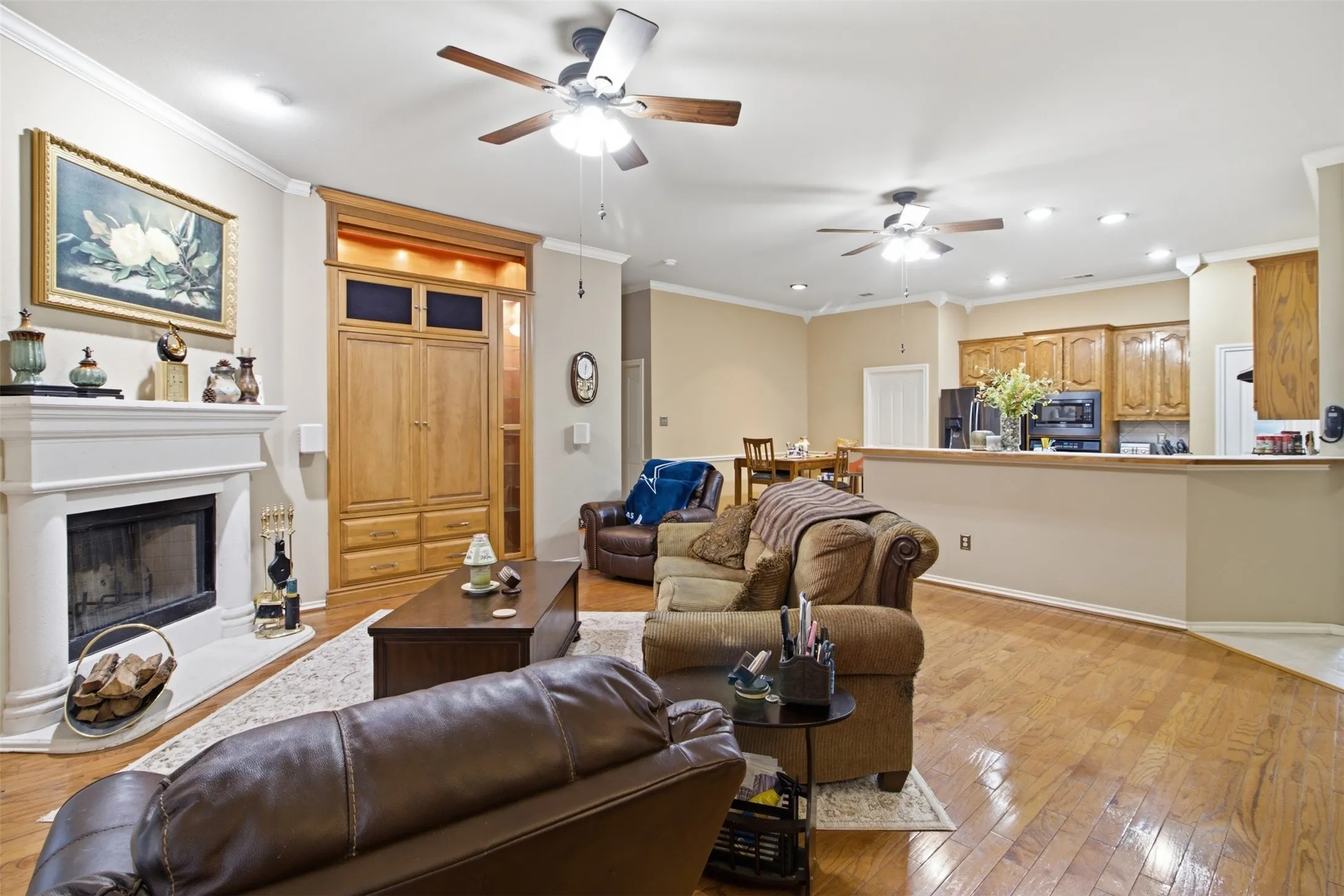 Hardwood flooring, crown molding, a built-in entertainment center, a fireplace with a mantel, and an open-concept kitchen with wood-finish cabinetry