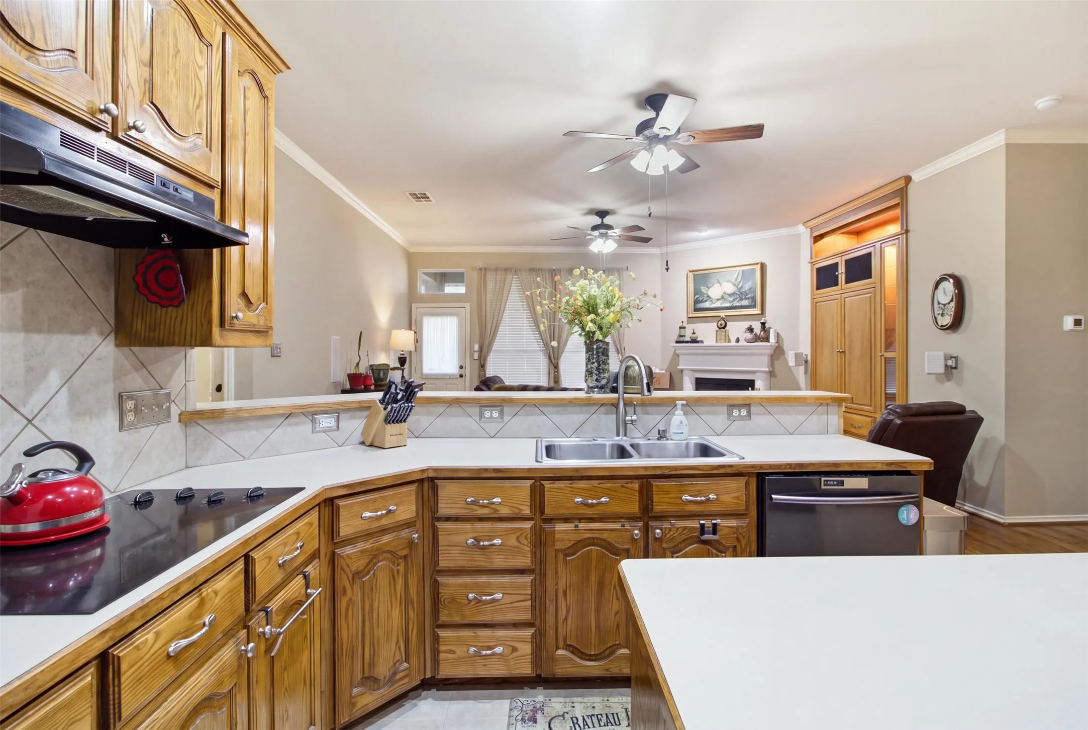 Wood-finish cabinetry, built-in electric cooktop, stainless steel double basin sink, tile backsplash, and overhead range hood