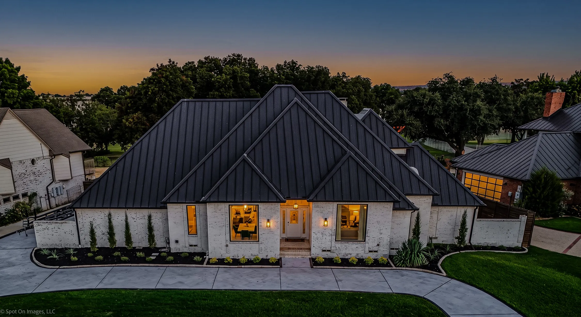 White brick exterior with a dark metal standing seam roof, featuring a concrete driveway, manicured landscaping, and exterior lighting fixtures