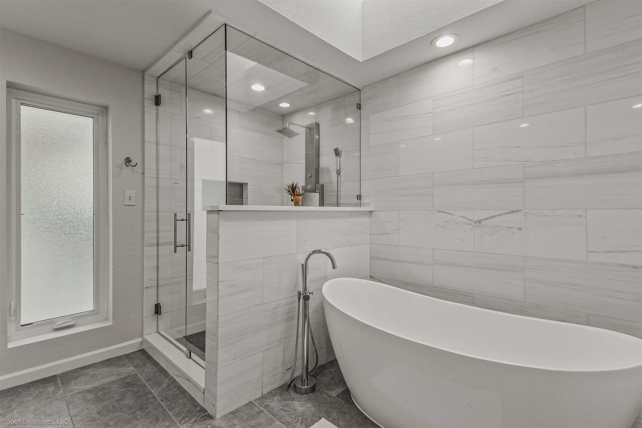Freestanding soaking tub with floor-mounted faucet, walk-in shower with glass enclosure, integrated shower shelf, and large format tiled walls
