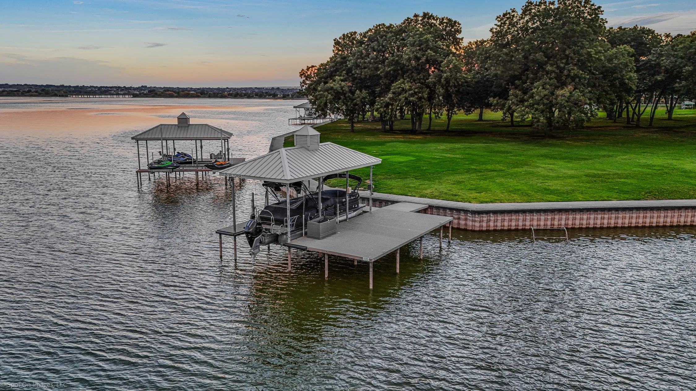 Waterfront property featuring two covered boat docks, an extensive green lawn, mature trees, and a lake retaining wall