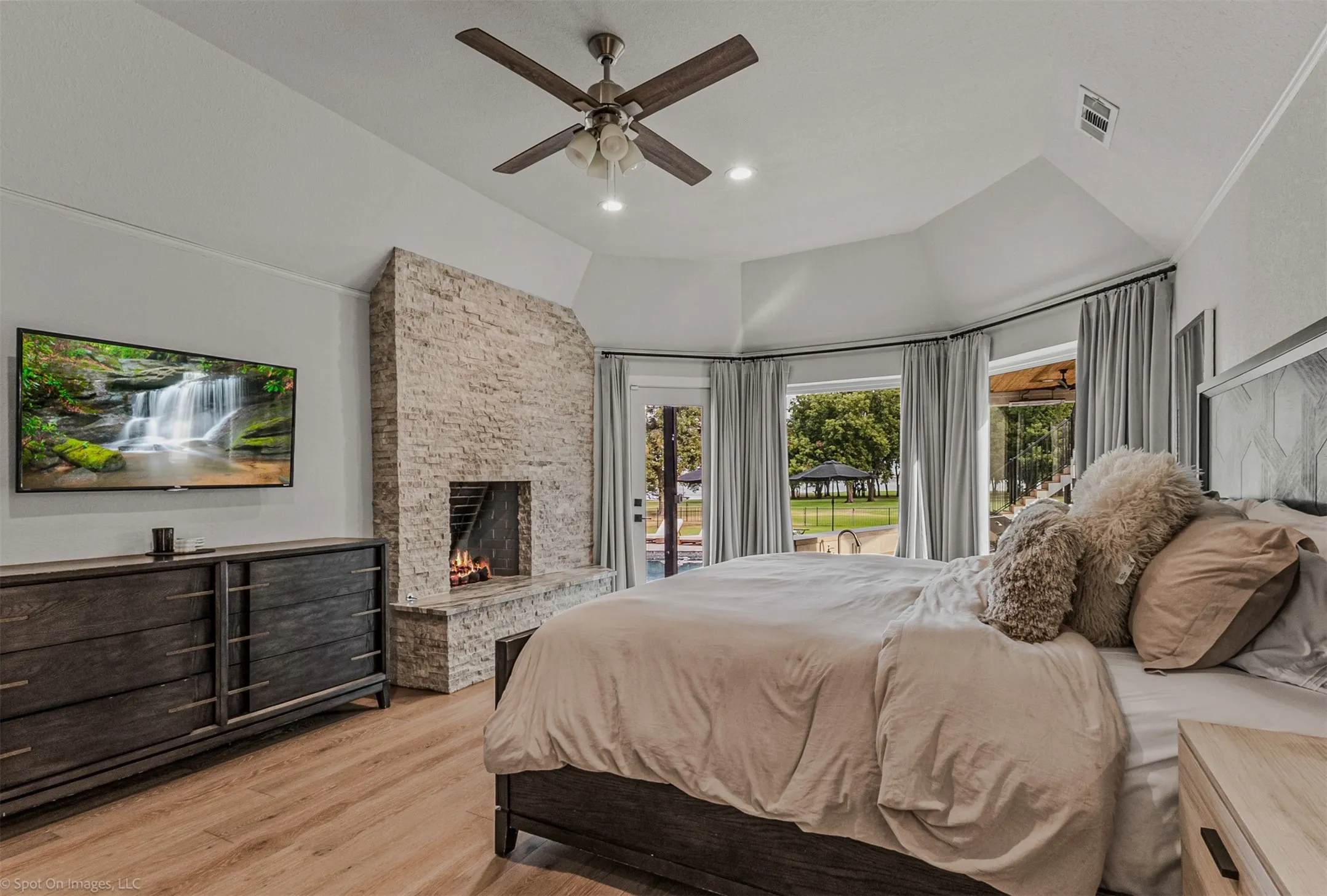Vaulted ceiling with a ceiling fan, stone-clad fireplace, wood-finish flooring, and multiple windows