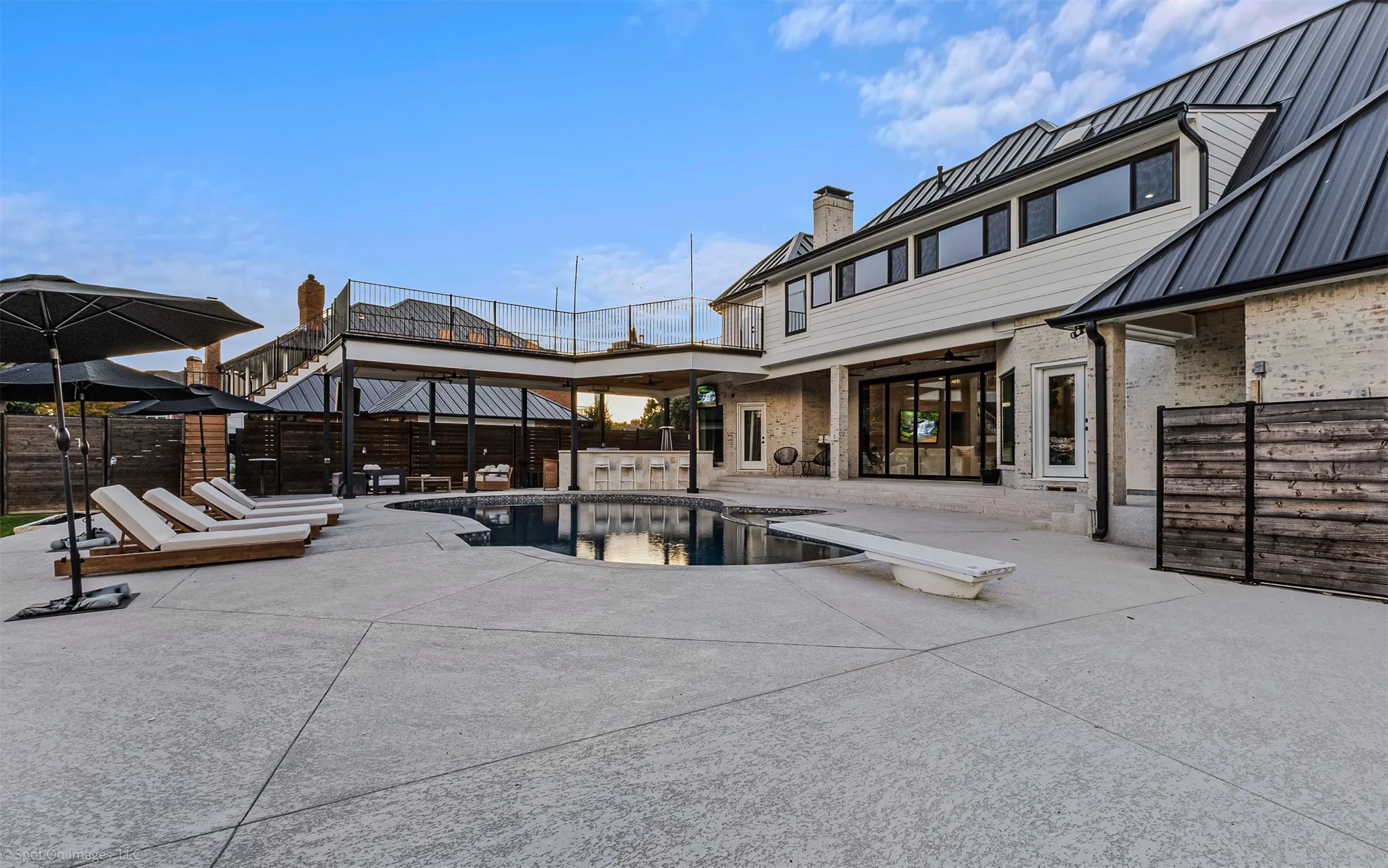 Expansive concrete patio with a built-in swimming pool, integrated diving board, and an outdoor kitchen area under a covered structure