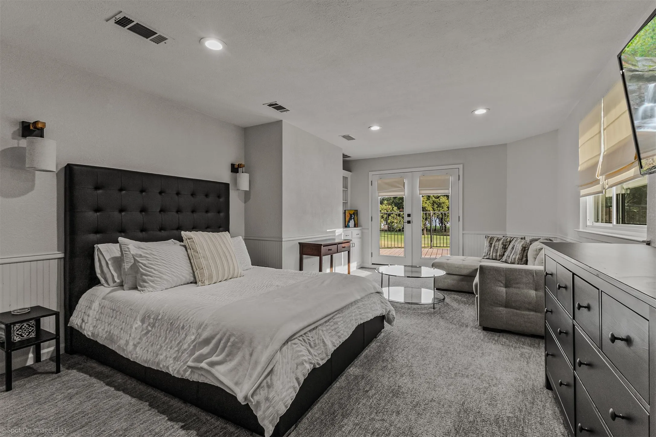 Spacious primary bedroom featuring a tufted black upholstered bed frame, recessed ceiling lighting, and dual glass doors opening to outdoor space