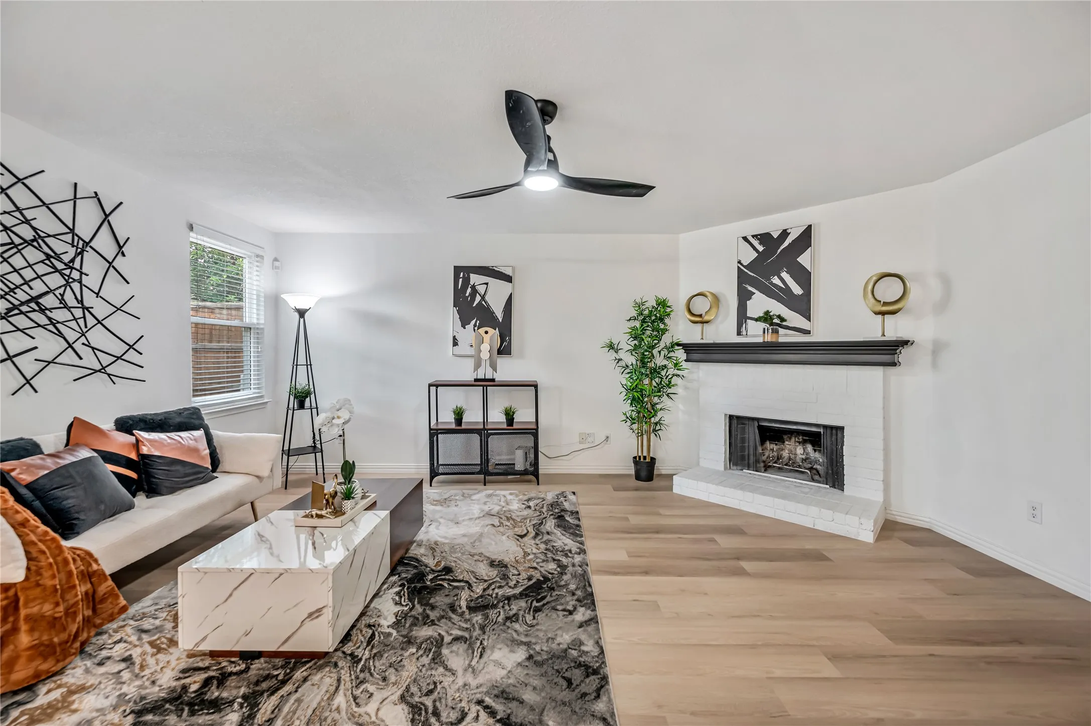 Light wood-finish flooring, white painted brick fireplace with black mantel, white walls, and a black ceiling fan