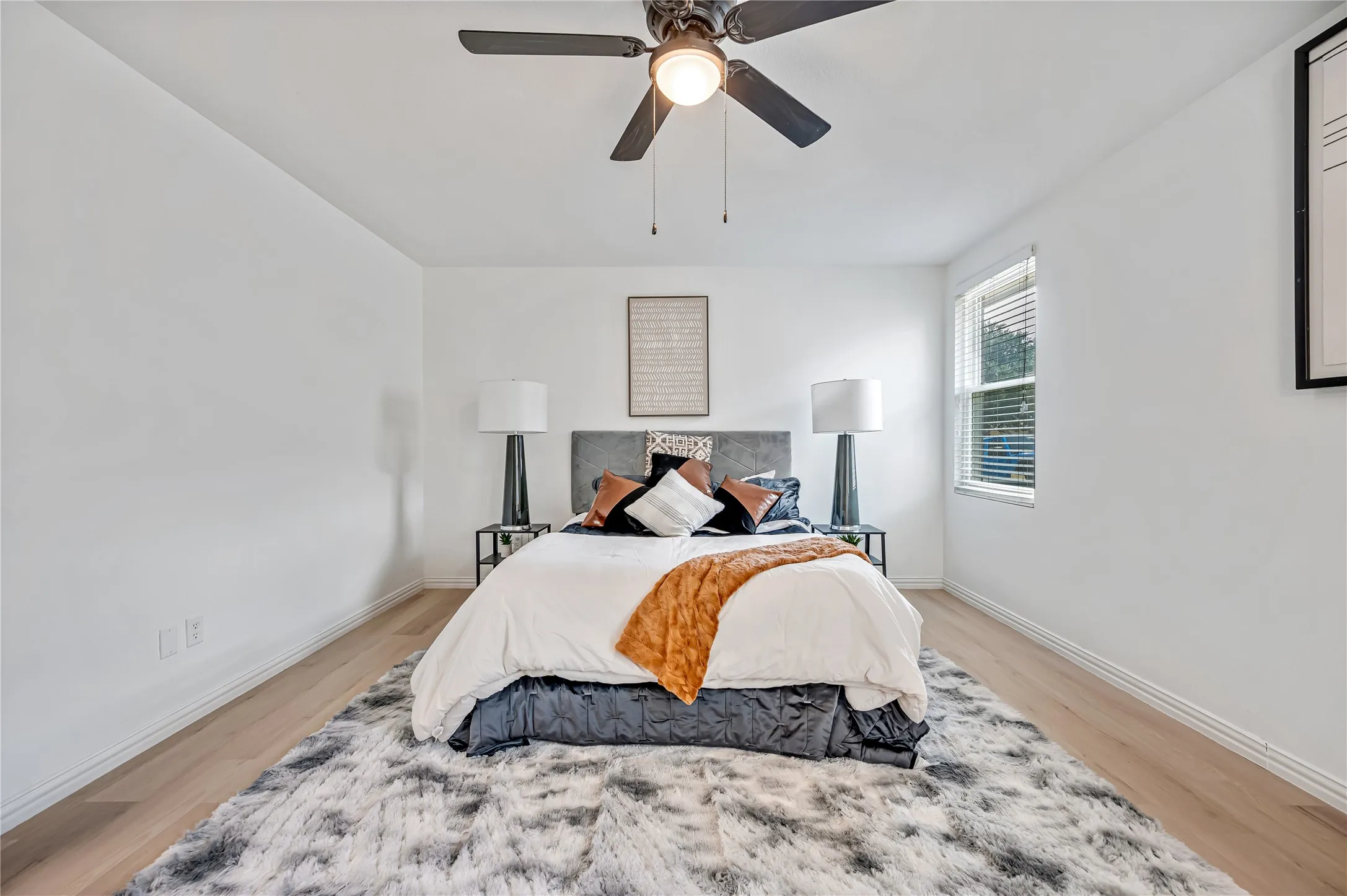 Light wood-finish flooring, white walls, black ceiling fan with integrated lighting, and a window with blinds
