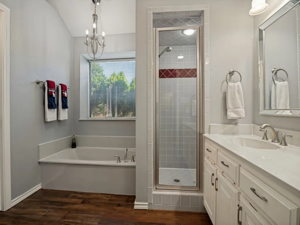 Spacious bathroom featuring a built-in soaking tub, separate glass-enclosed shower with tile surround, wood-finish flooring, a vanity with an integrated sink and white countertop, and a reflective framed mirror