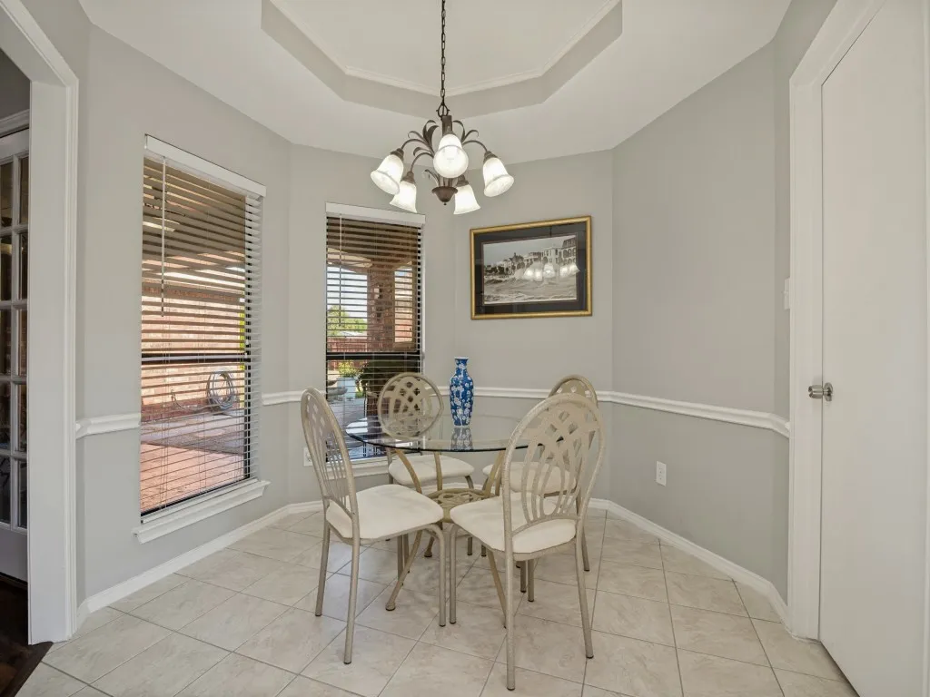 Recessed tray ceiling with crown molding, tiled flooring, two windows with blinds, chair rail molding, and a chandelier