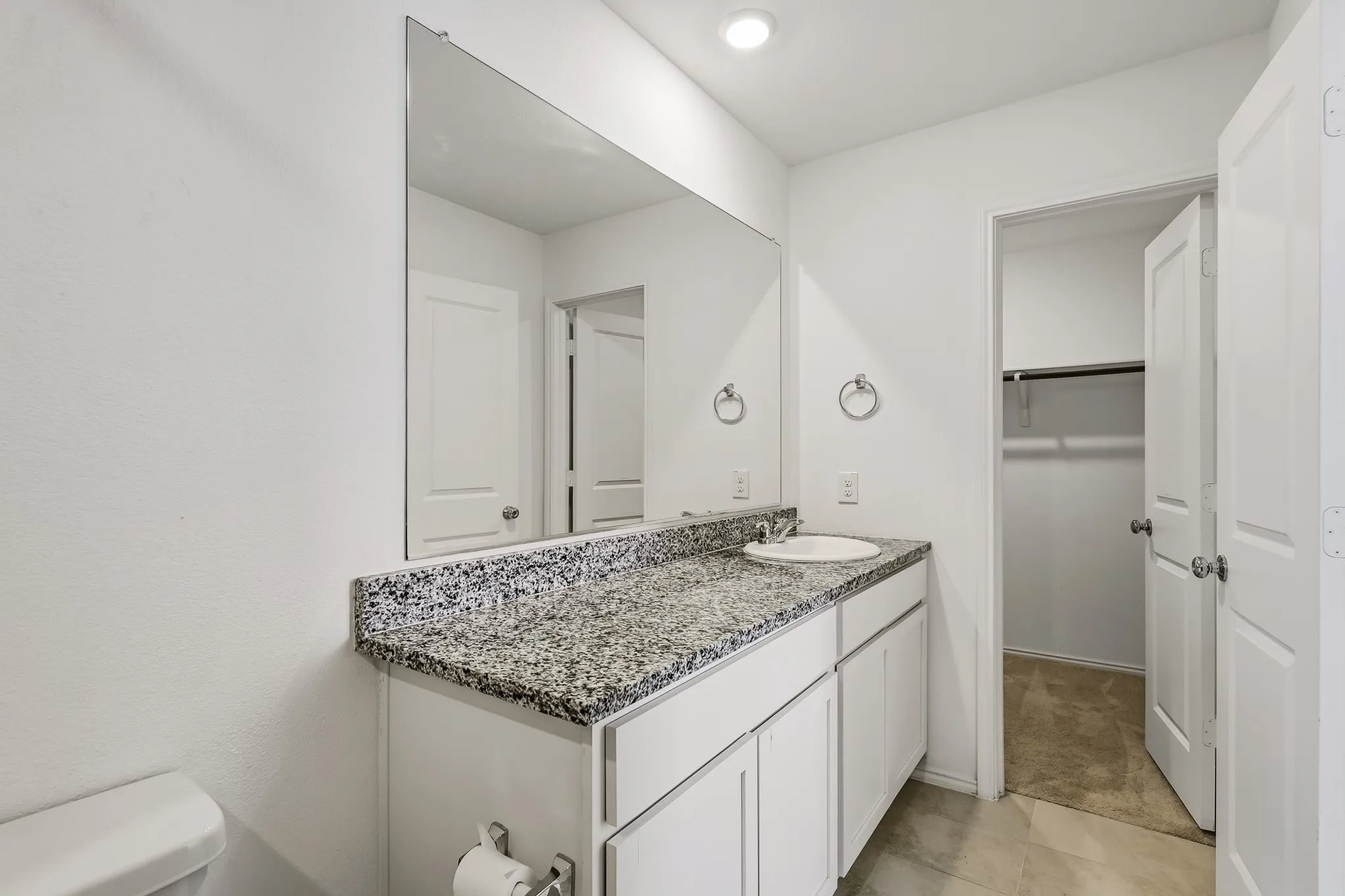 Vanity with a speckled countertop and integrated sink, white cabinetry, large wall-mounted mirror, and tile flooring