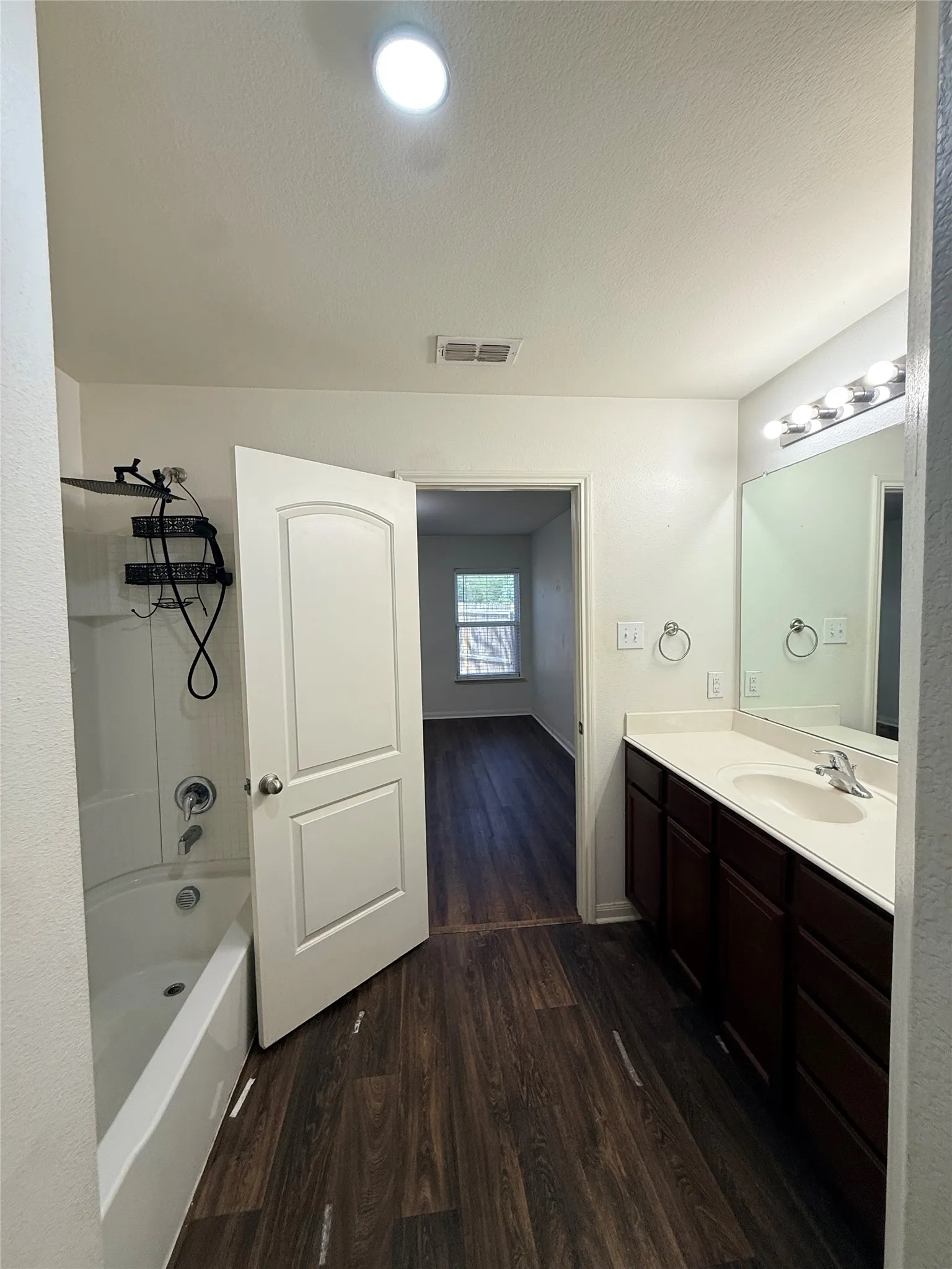 Wood-finish flooring, white vanity with integrated sink, chrome faucet, wall-mounted mirror with vanity lighting, recessed ceiling light