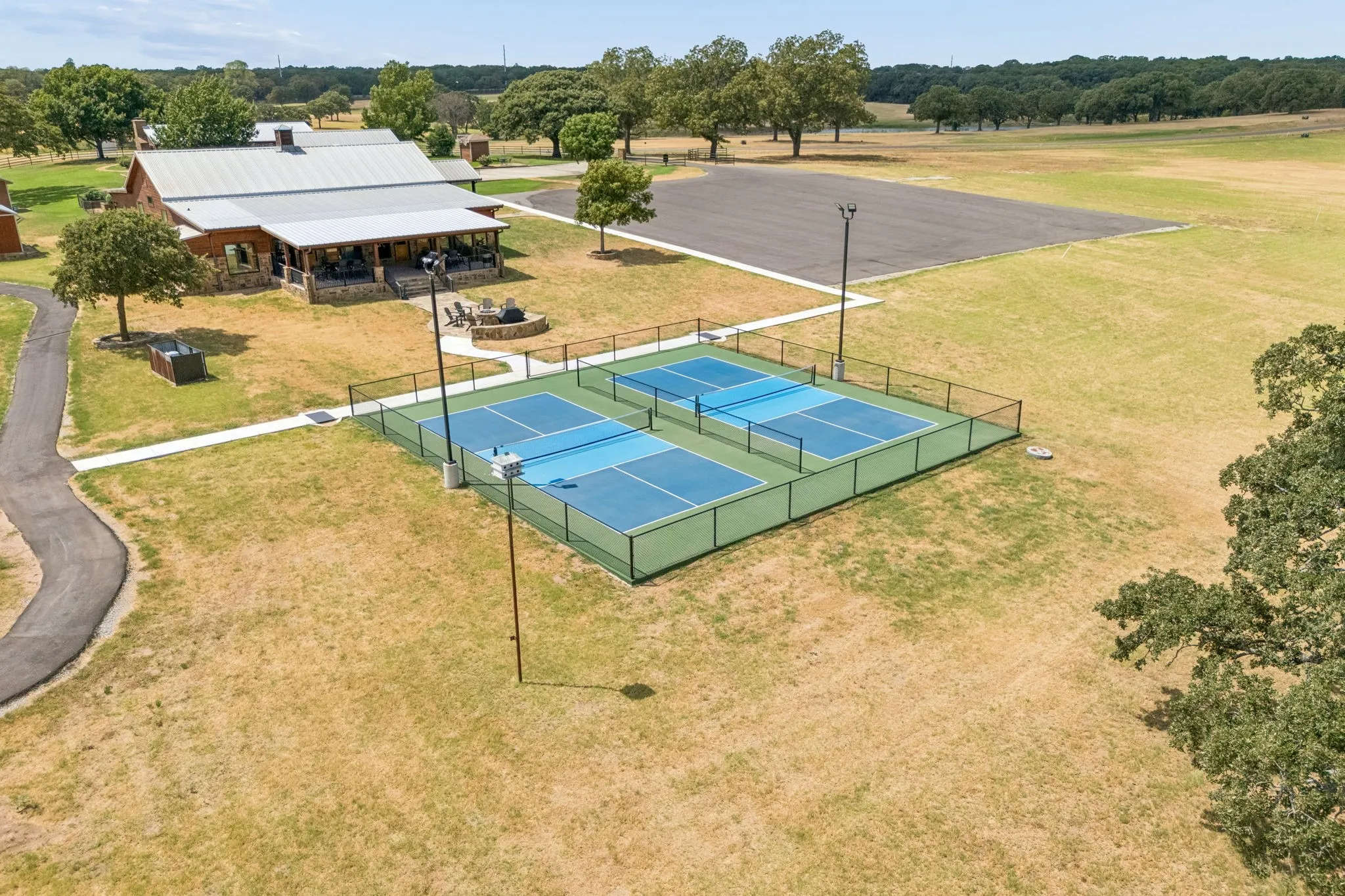 Two fenced pickleball courts, a large building with an expansive covered porch, a paved parking area, and mature trees across the property