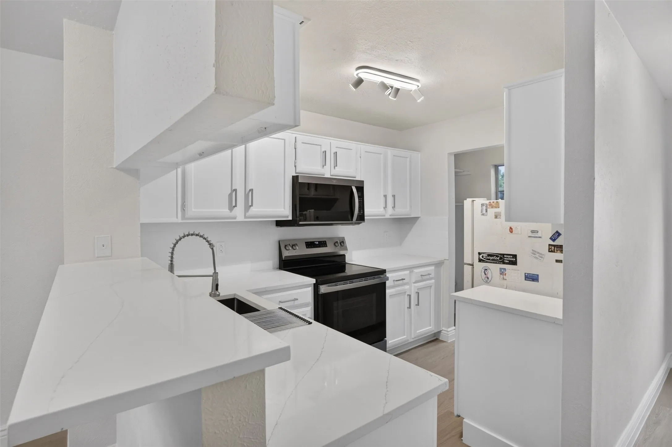 White shaker-style cabinetry, stainless steel sink with gooseneck faucet, light-toned countertops, contemporary track lighting, and wood-finish flooring