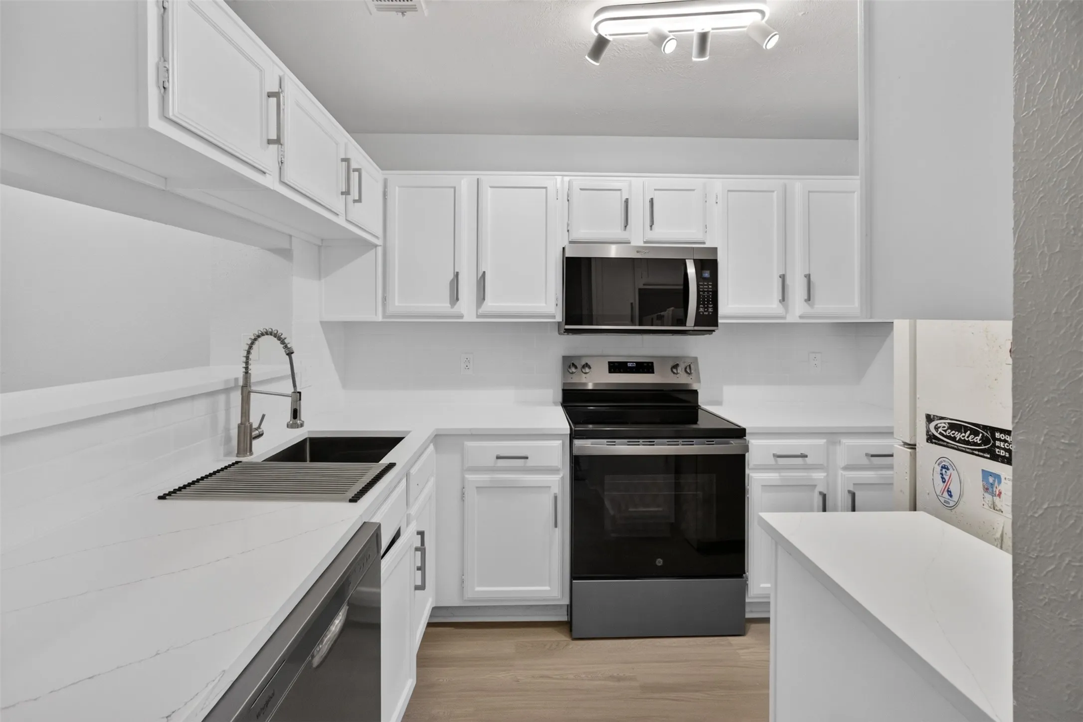 White cabinetry with brushed nickel hardware, stainless steel appliances, dual basin sink with gooseneck faucet, light wood-finish flooring, and white countertops
