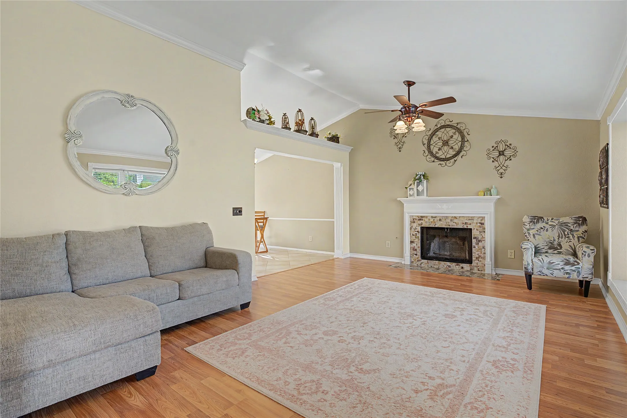 Vaulted ceilings and wood-finish flooring enhance this living space, featuring a stone-surround fireplace with a classic mantel, an overhead ceiling fan with integrated lighting, and architectural trim details