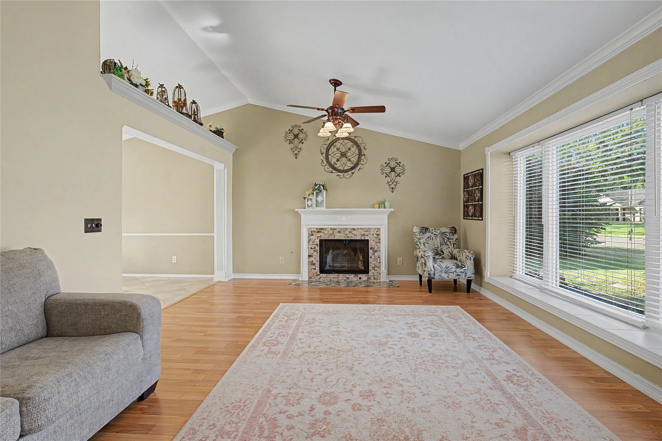 Living room featuring vaulted ceilings, wood-finish flooring, a tiled fireplace with a mantel, a ceiling fan with integrated lighting, and a large bay window