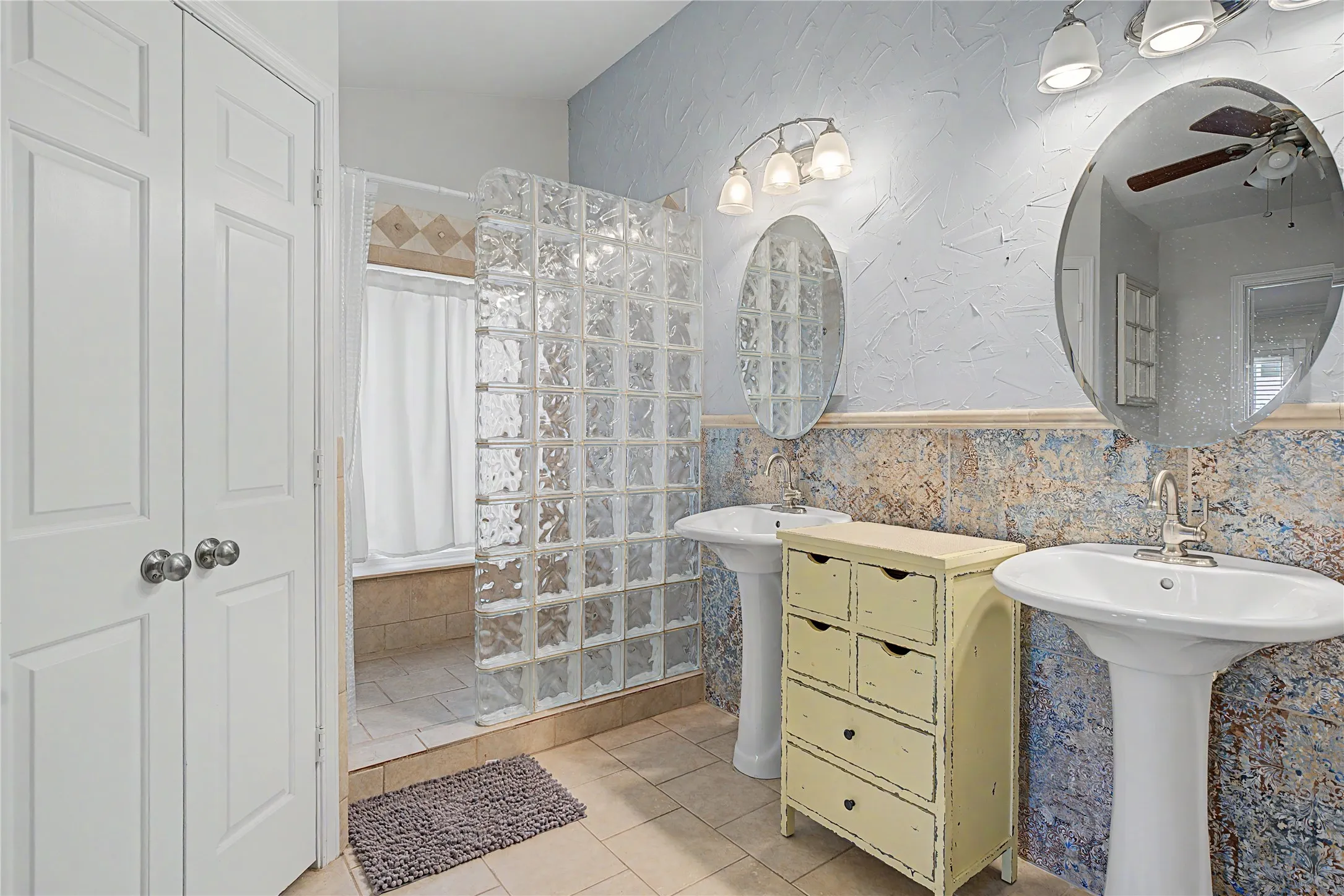 Dual pedestal sinks, glass block shower enclosure, tiled wainscoting, textured wall finish, and two-panel closet doors